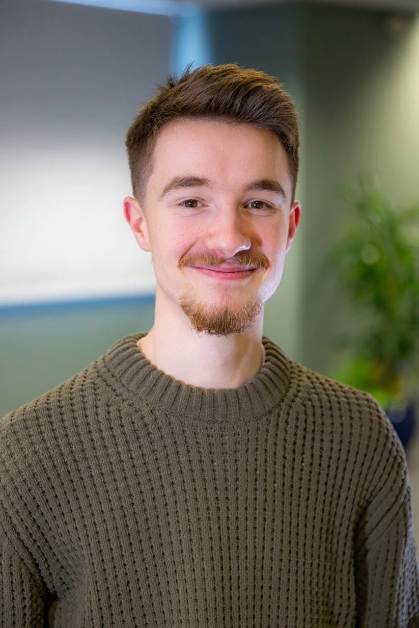 A young man with brown hair, a mustache, and beard, smiling, wearing a green knitted sweater, in an indoor setting with a blurred background of a window and a green plant.