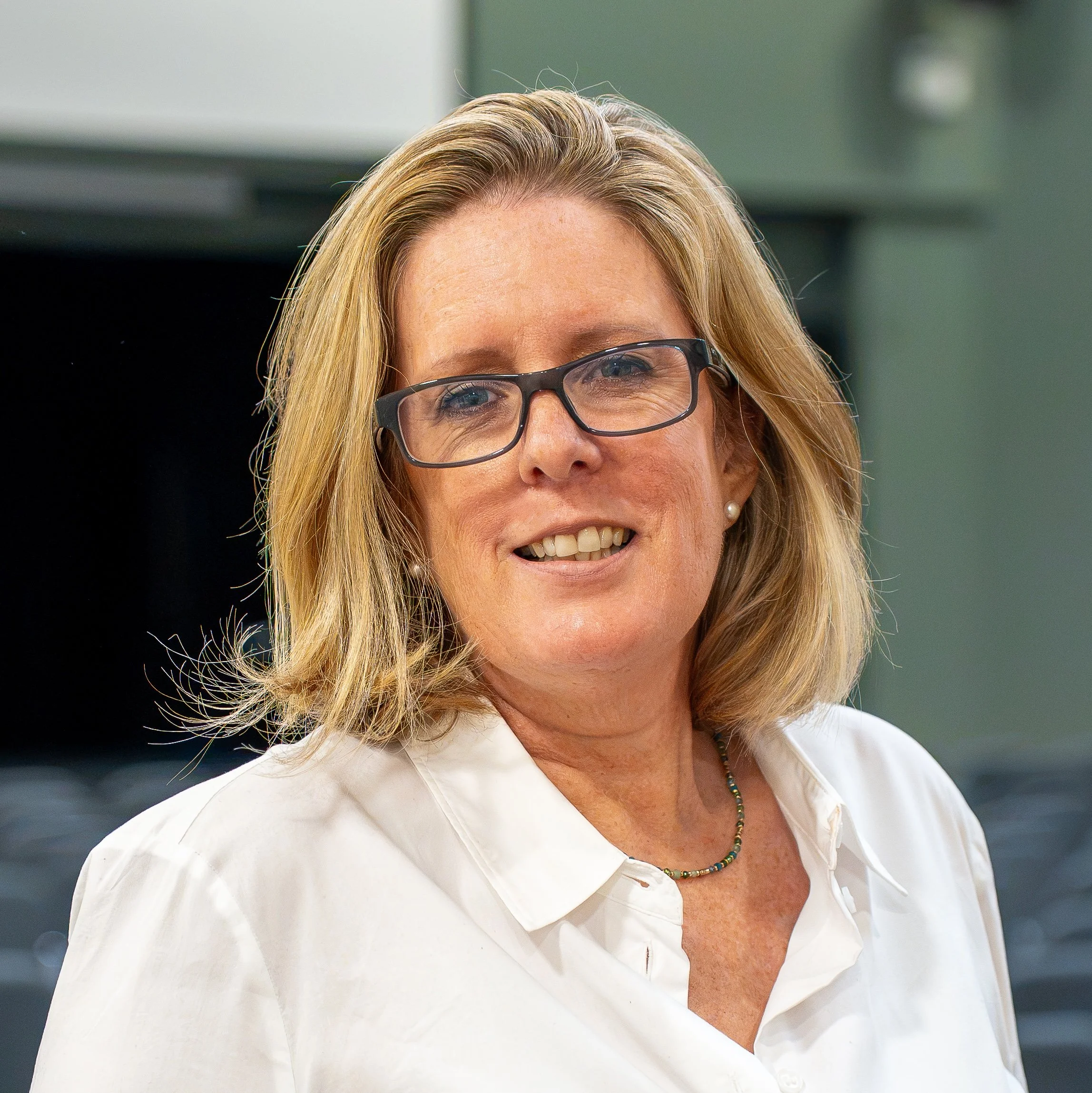Close-up of a woman with blonde hair, wearing glasses, a white blouse, and a beaded necklace, smiling outdoors.