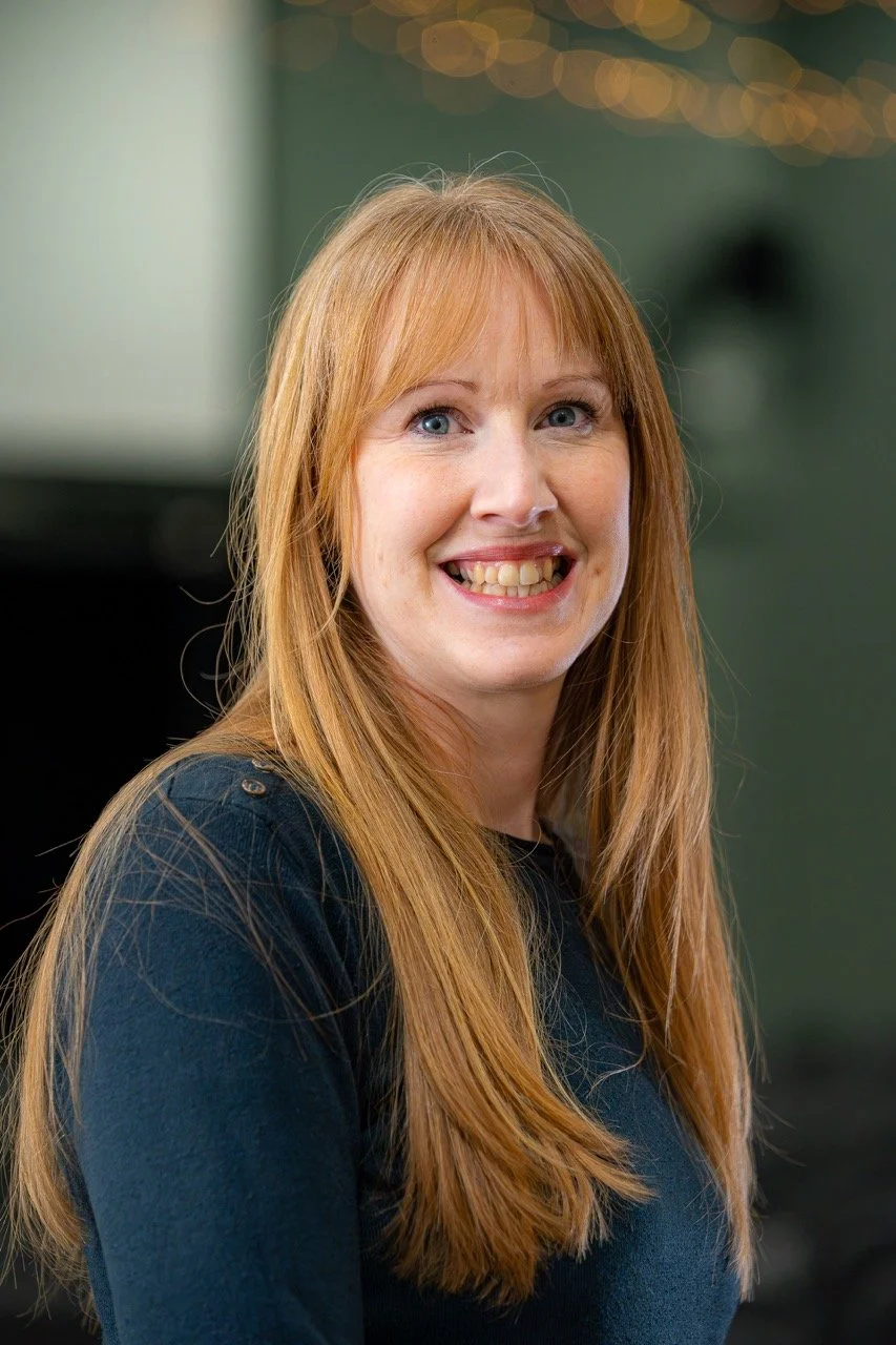 A woman with long red hair smiling at the camera in an indoor setting.