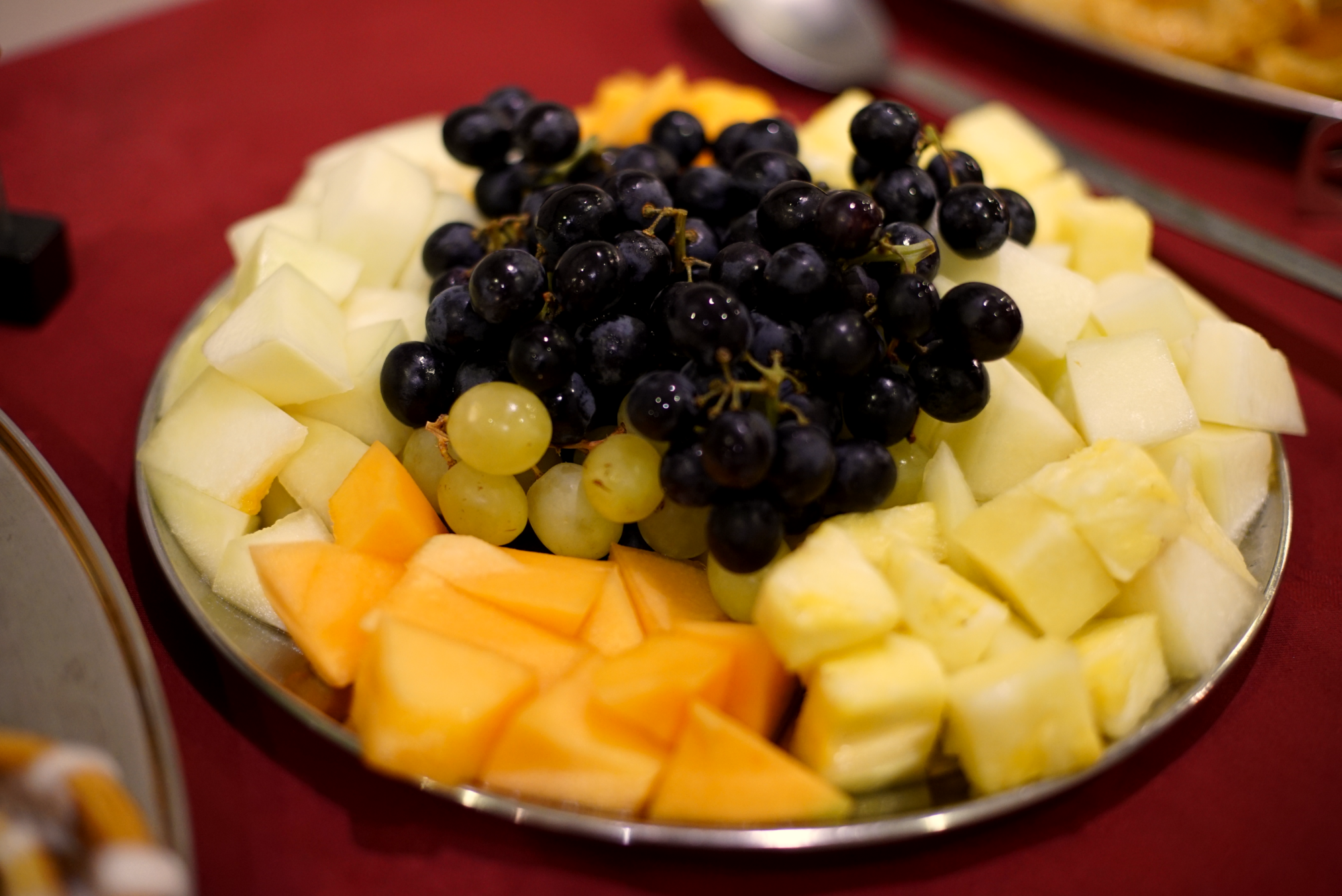 A round metal tray filled with assorted fresh fruit. It includes grapes, honeydew melon, cantaloupe, pineapple, and cantaloupe.