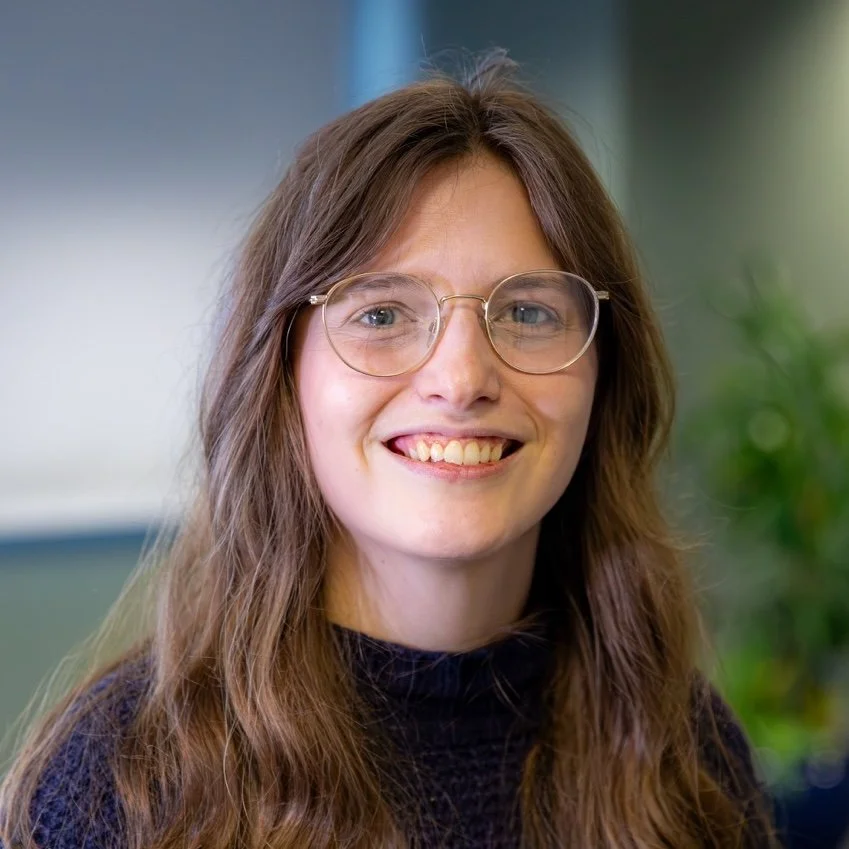 A young woman with long brown hair, glasses, and a black turtleneck smiling at the camera.