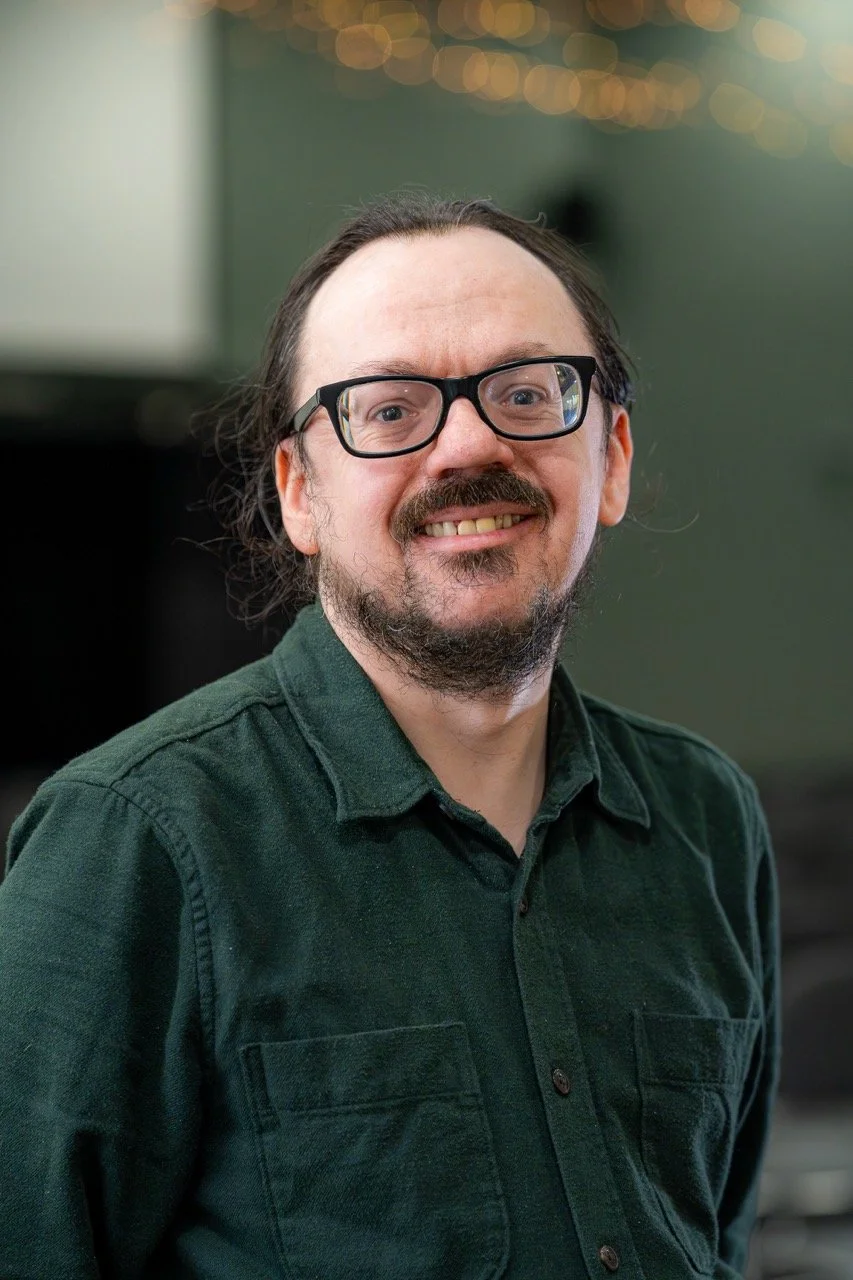 A man with glasses and a beard, smiling, wearing a dark green shirt, in an indoor setting with blurred lights in the background.