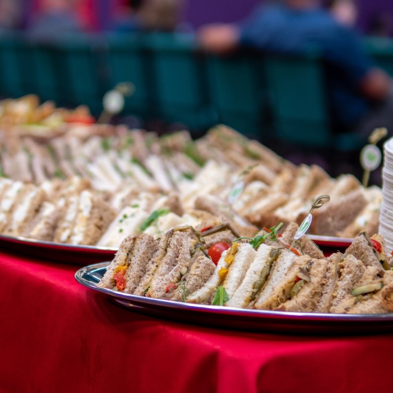 Assorted finger sandwiches on a platter with a red tablecloth, with blurred people in the background. Taken at a Norwich event using King's Catering.