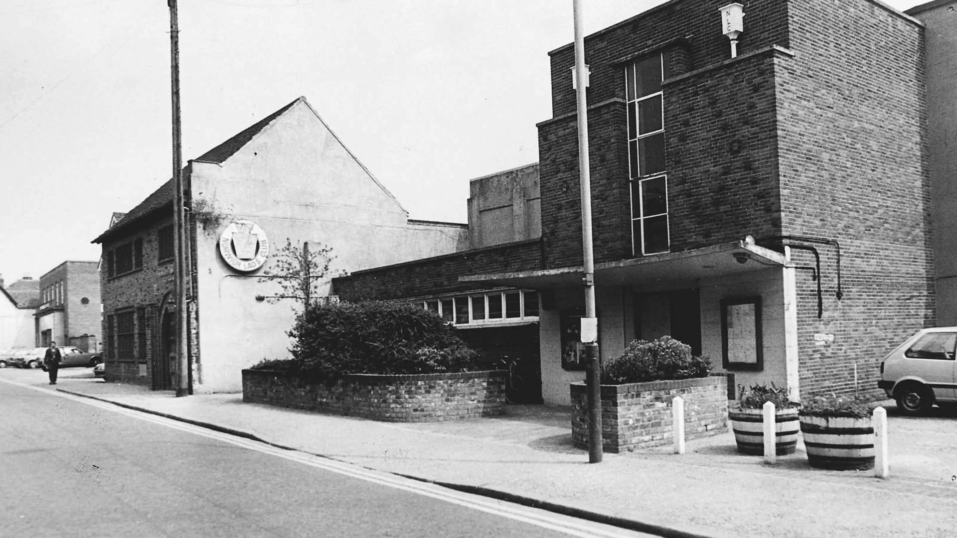 Black-and-white photo of King Street with a man walking, showing what the King's Centre originally looked like with a circular sign, bushes in brick planters, and modern brick buildings in the background.