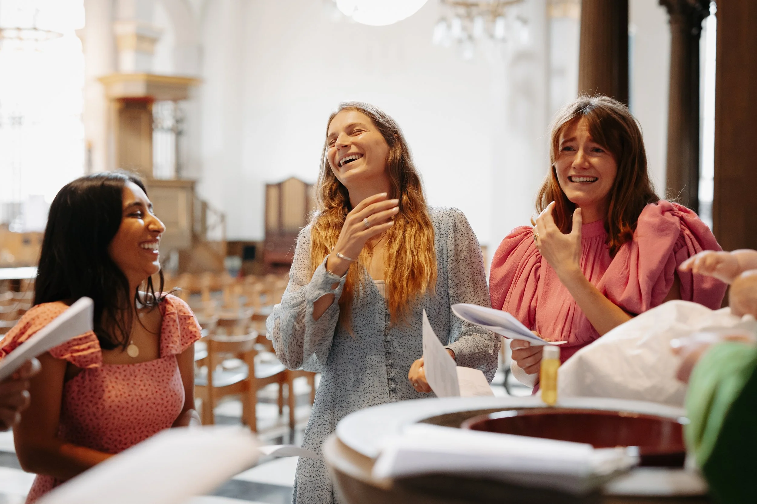 Three women happily chatting near a food counter in a brightly lit restaurant or market. One woman in a pink dress, another in a white dress, and the third in a red dress are smiling and holding papers or menus.