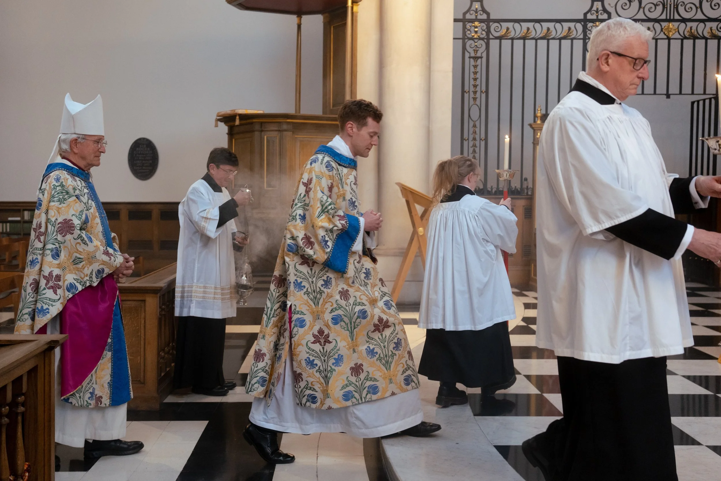 Clergy members dressed in religious vestments are participating in a church service. They are standing in a row with some holding incense and candles, and their heads bowed in prayer or reflection inside a church with ornate black and gold railing and a black and white checkered floor.