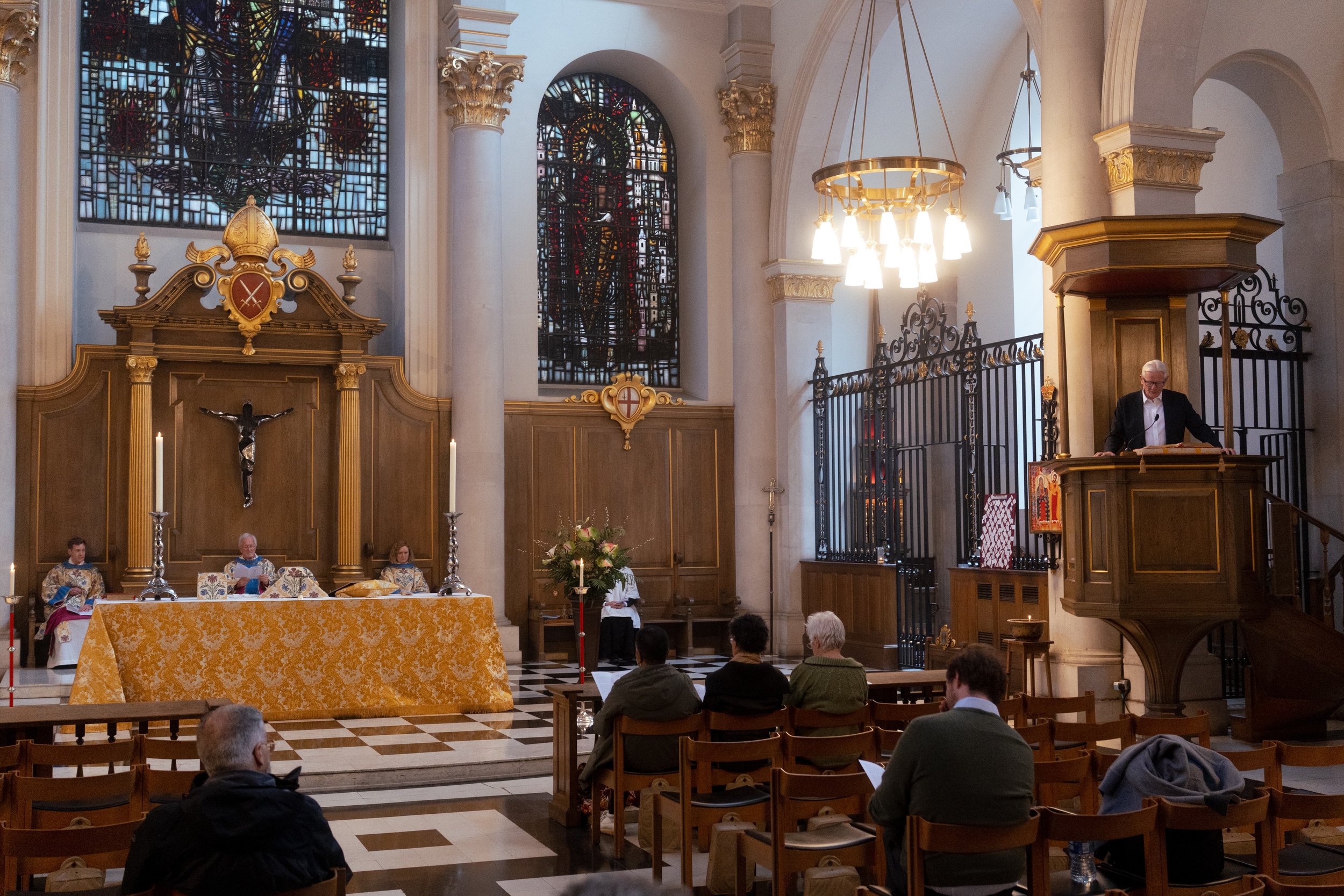 Inside a church with high ceilings, stained glass windows, and ornate gold and wood decorations. Several people are seated, some at an altar with candles and flowers, while a man stands at a pulpit delivering a sermon or reading. The altar has religious symbols and a crucifix.