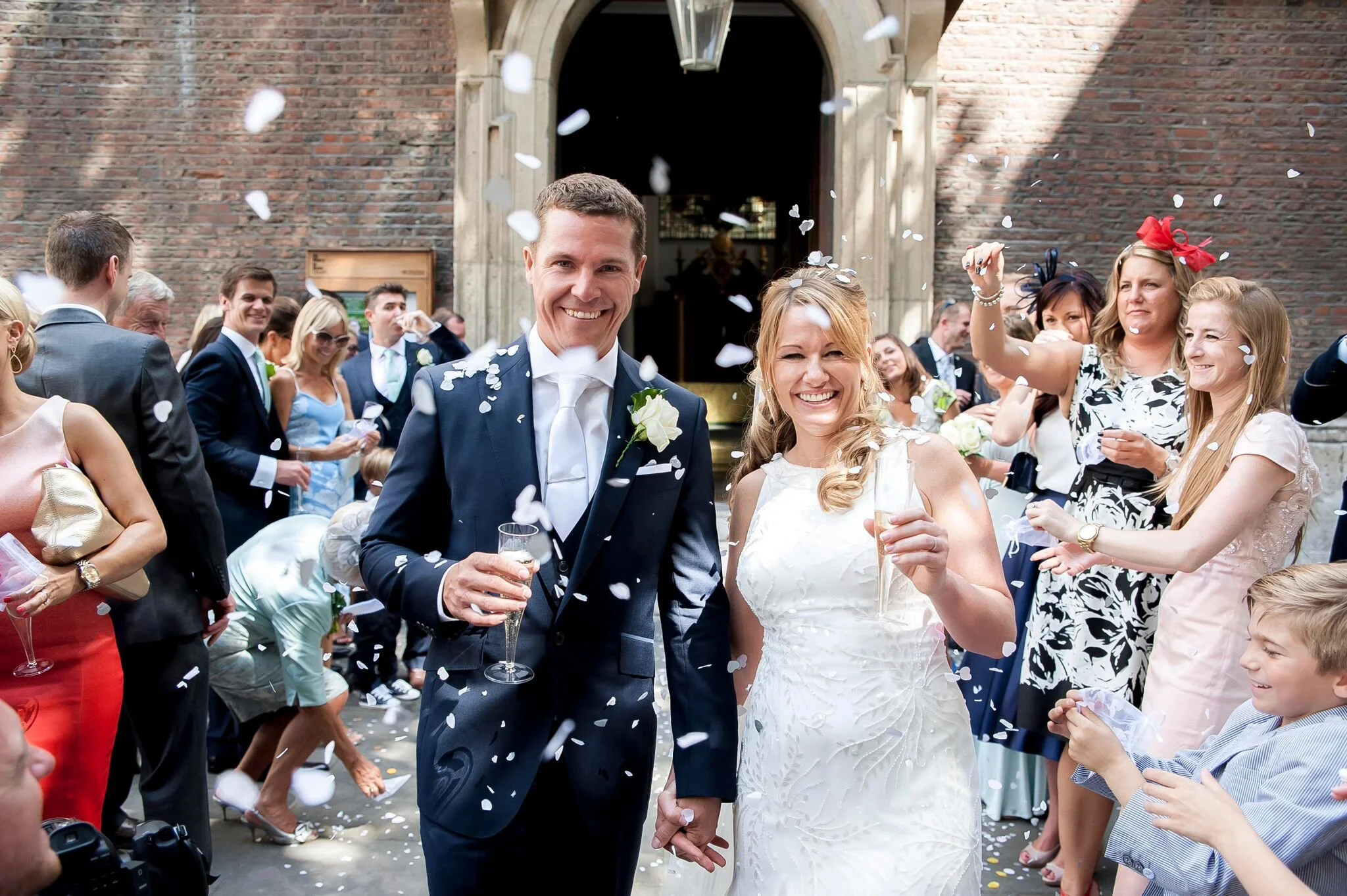 A bride and groom smiling and holding hands as they celebrate their wedding outside a church. Guests surround them, throwing white confetti.
