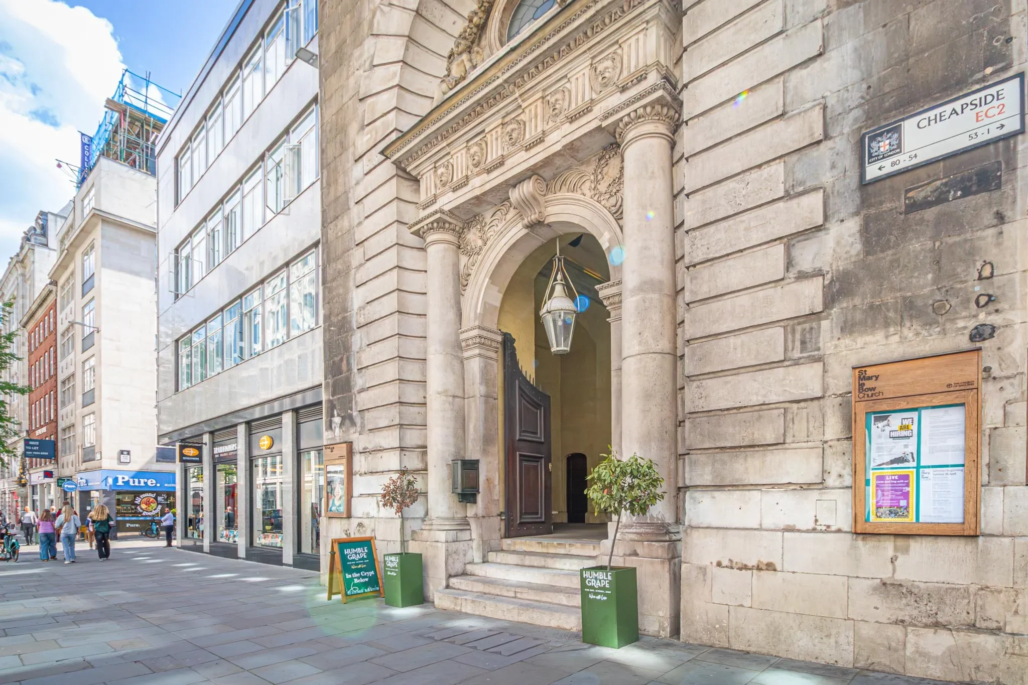 An urban street scene with a historic building featuring an arched entrance and decorative stonework, surrounded by modern buildings with large glass windows, pedestrians walking on the sidewalk, and street signs indicating nearby locations.