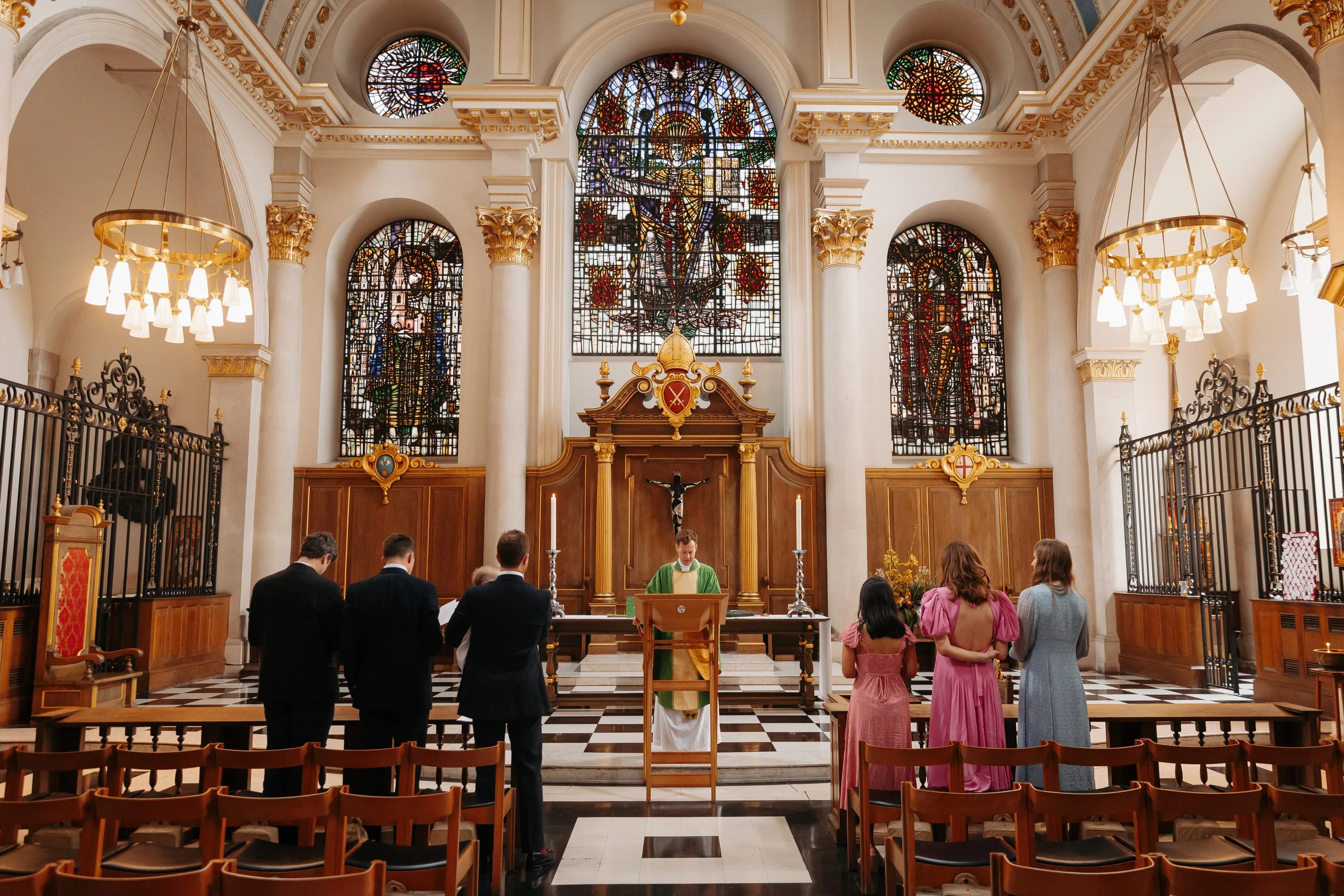 Inside a church with stained glass windows, an altar, and people participating in a religious ceremony, including a priest wearing green vestments.