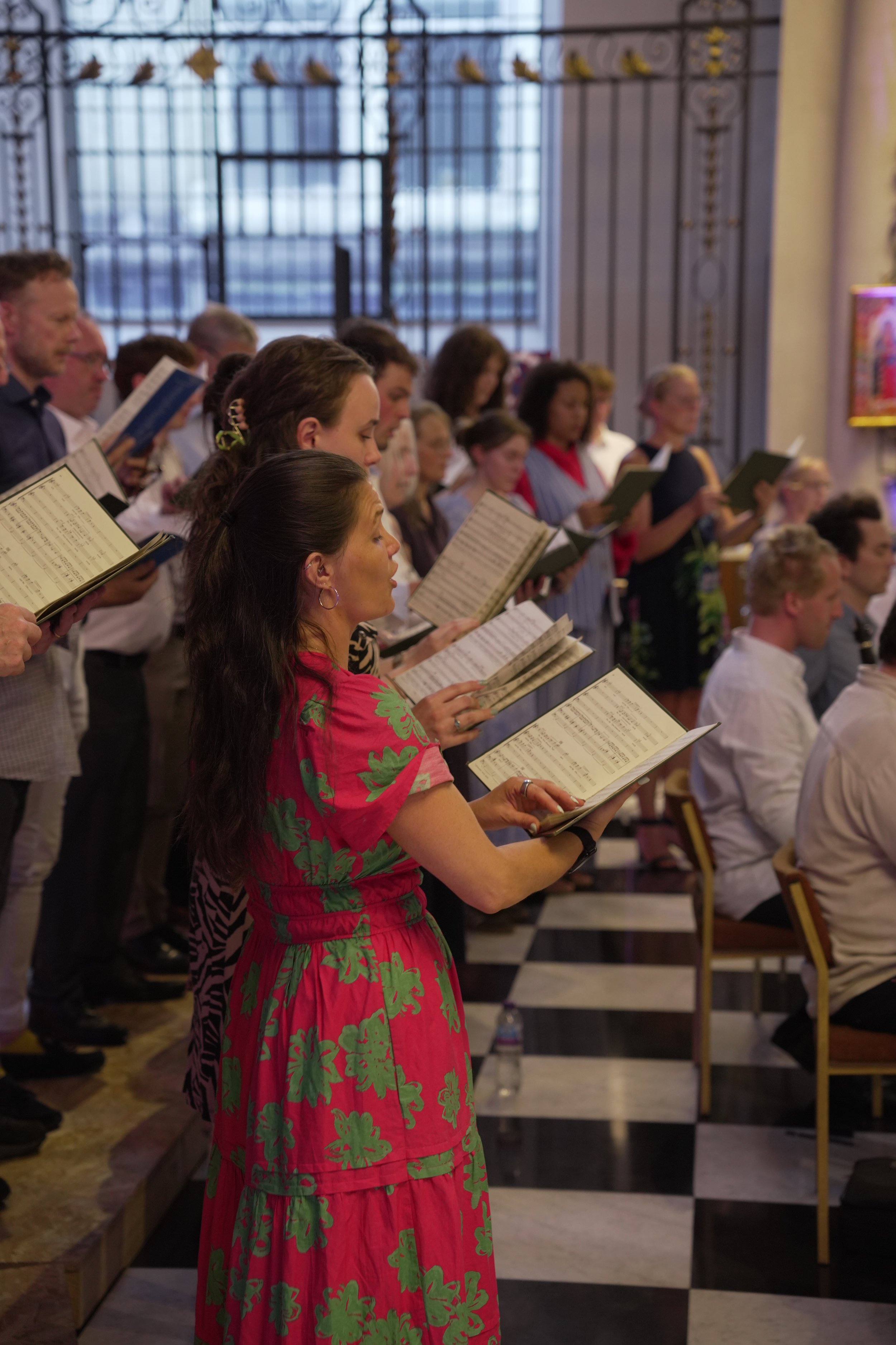 Group of people singing in a church or chapel, holding sheet music, with stained glass windows and a decorative iron gate in the background.