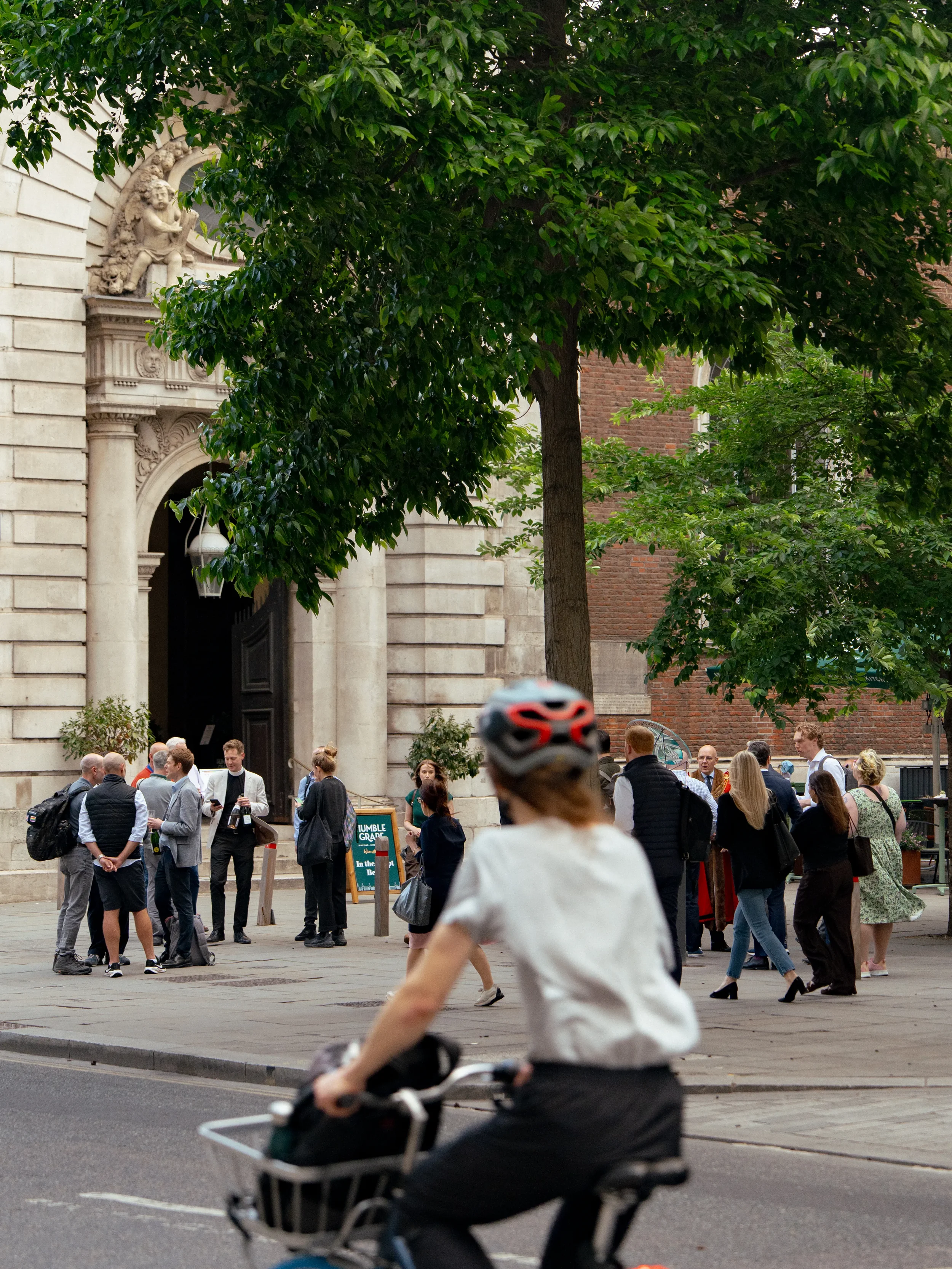 A crowd of people gathered outside a historic building with ornate stone architecture and sculptures. A person wearing a bicycle helmet is riding past in the foreground.