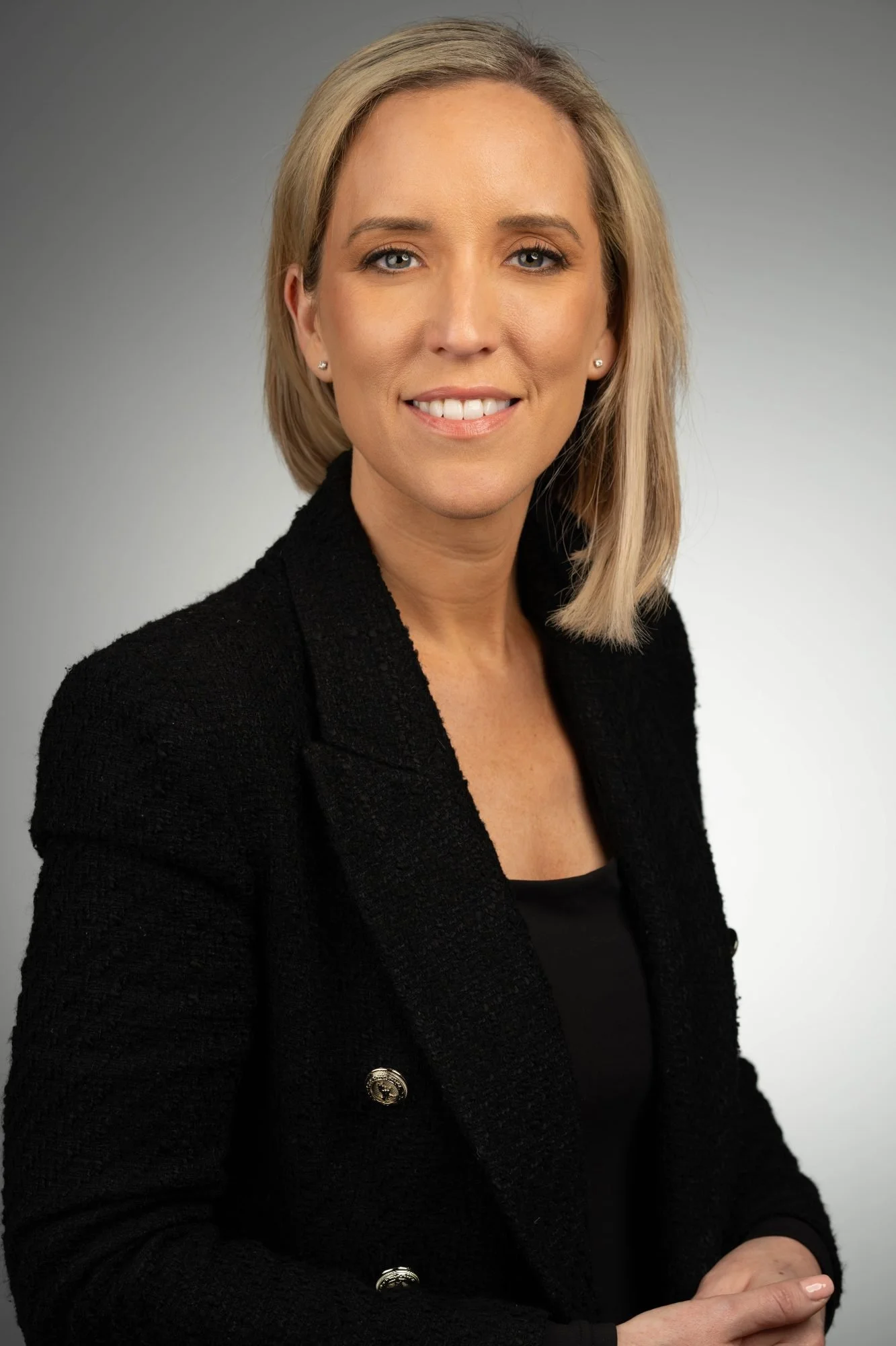 A woman with blonde hair, wearing a black textured blazer with silver buttons, smiling at the camera against a neutral gray background.