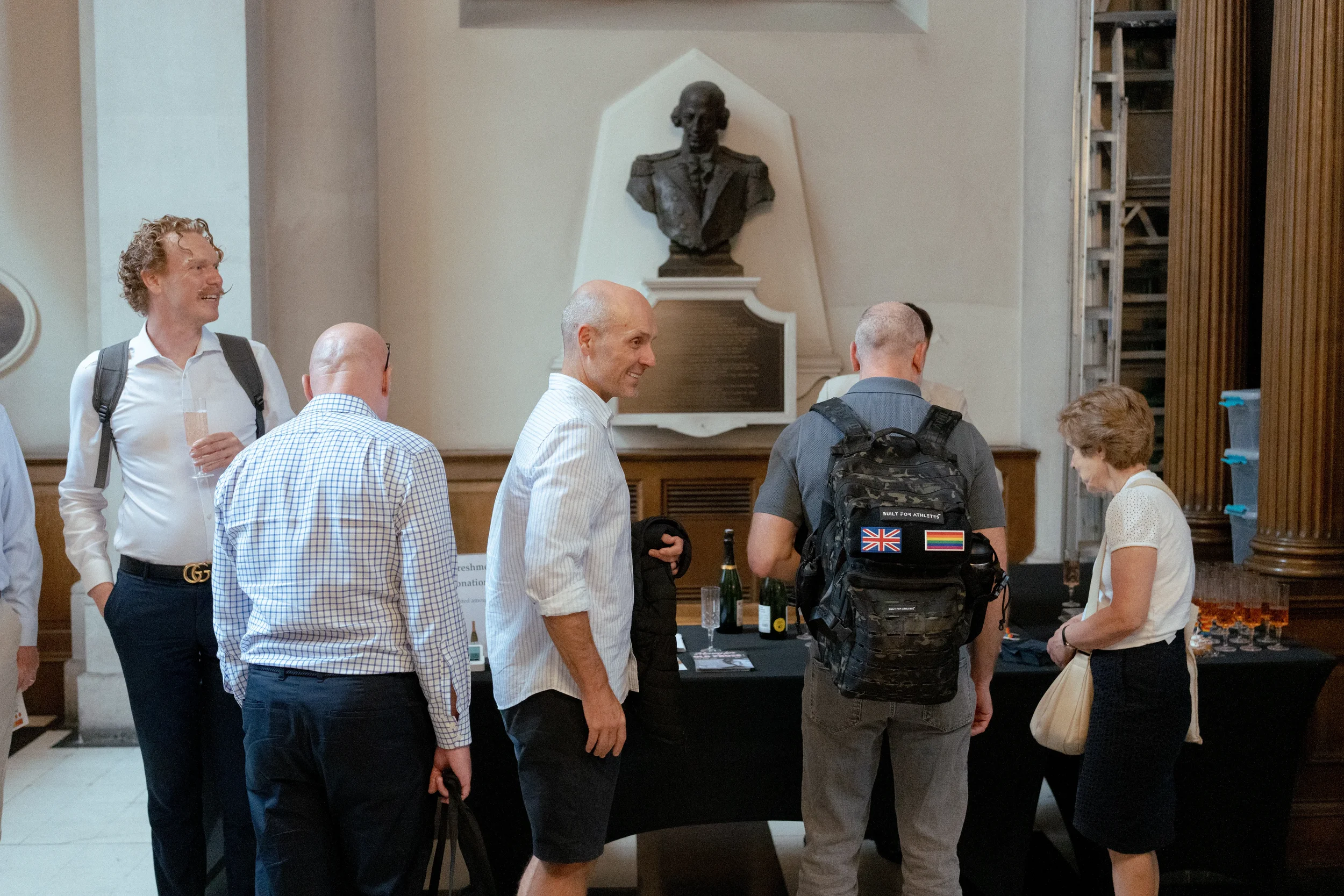 Group of people engaging in conversation at an indoor event, with a bust sculpture on a wall plaque in the background.