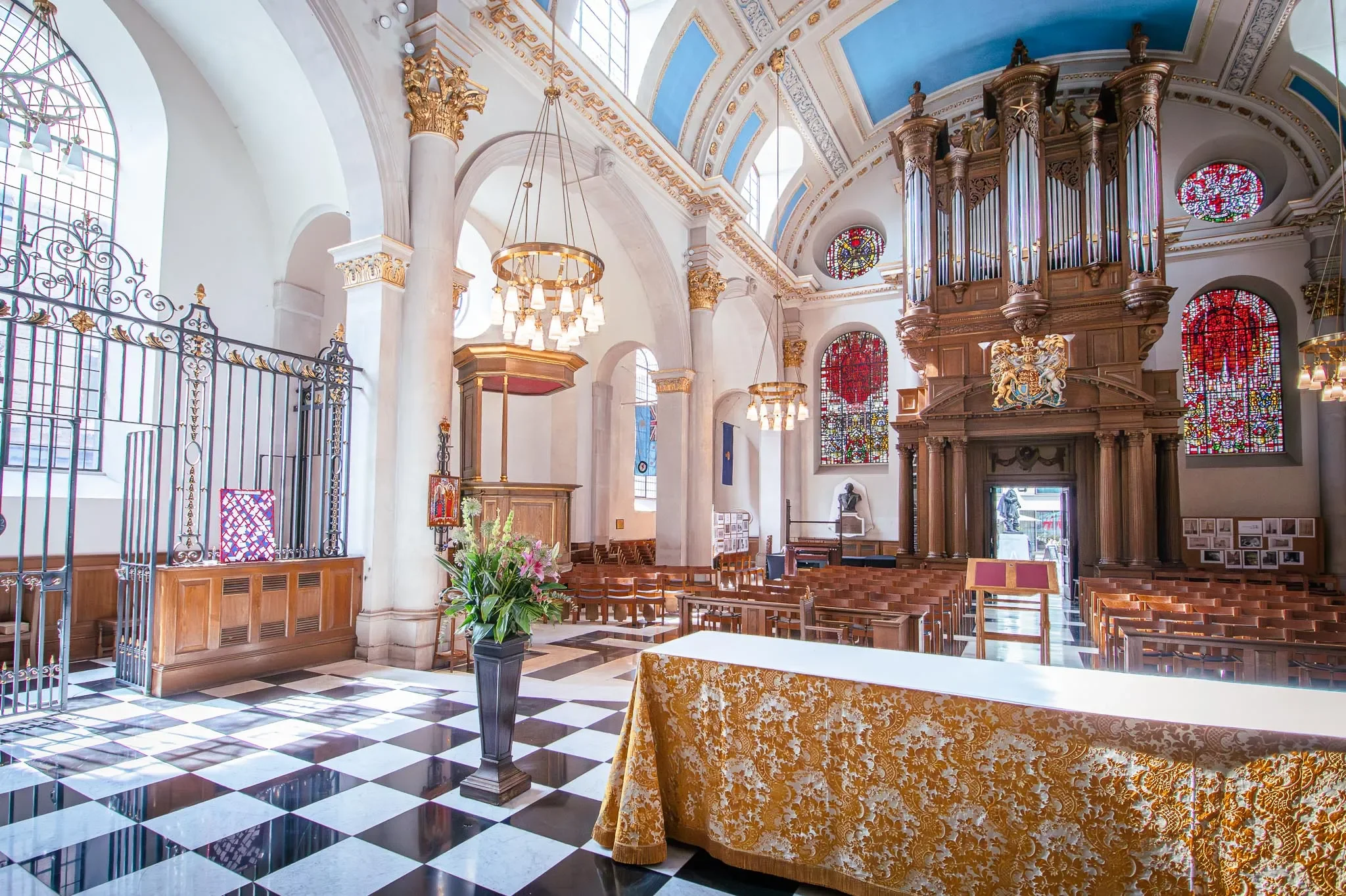Interior of a church with high vaulted ceilings painted blue and decorated with gold accents. Large stained-glass windows with red designs, ornate wooden organ pipes, a table with a gold-patterned cloth, a vases of flowers, and gold chandeliers hanging from the ceiling.