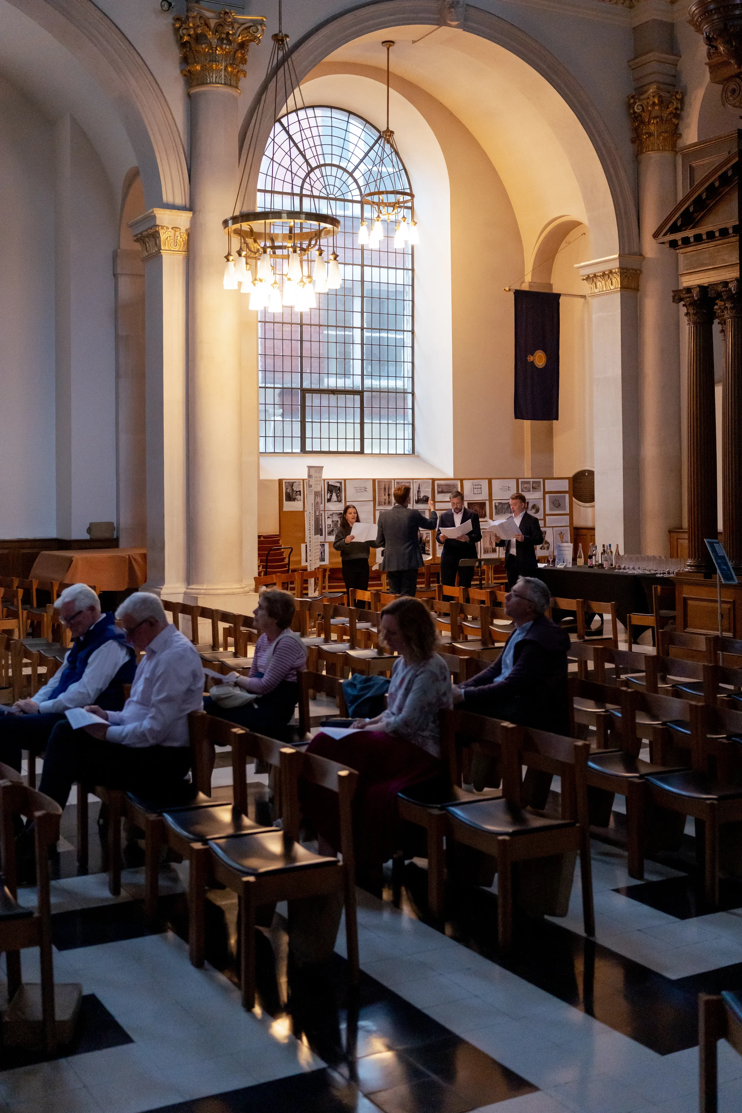 People attending a formal event in a church with high ceilings, large stained-glass window, ornate columns, and a chandelier, seated on wooden chairs, with some viewing programs.