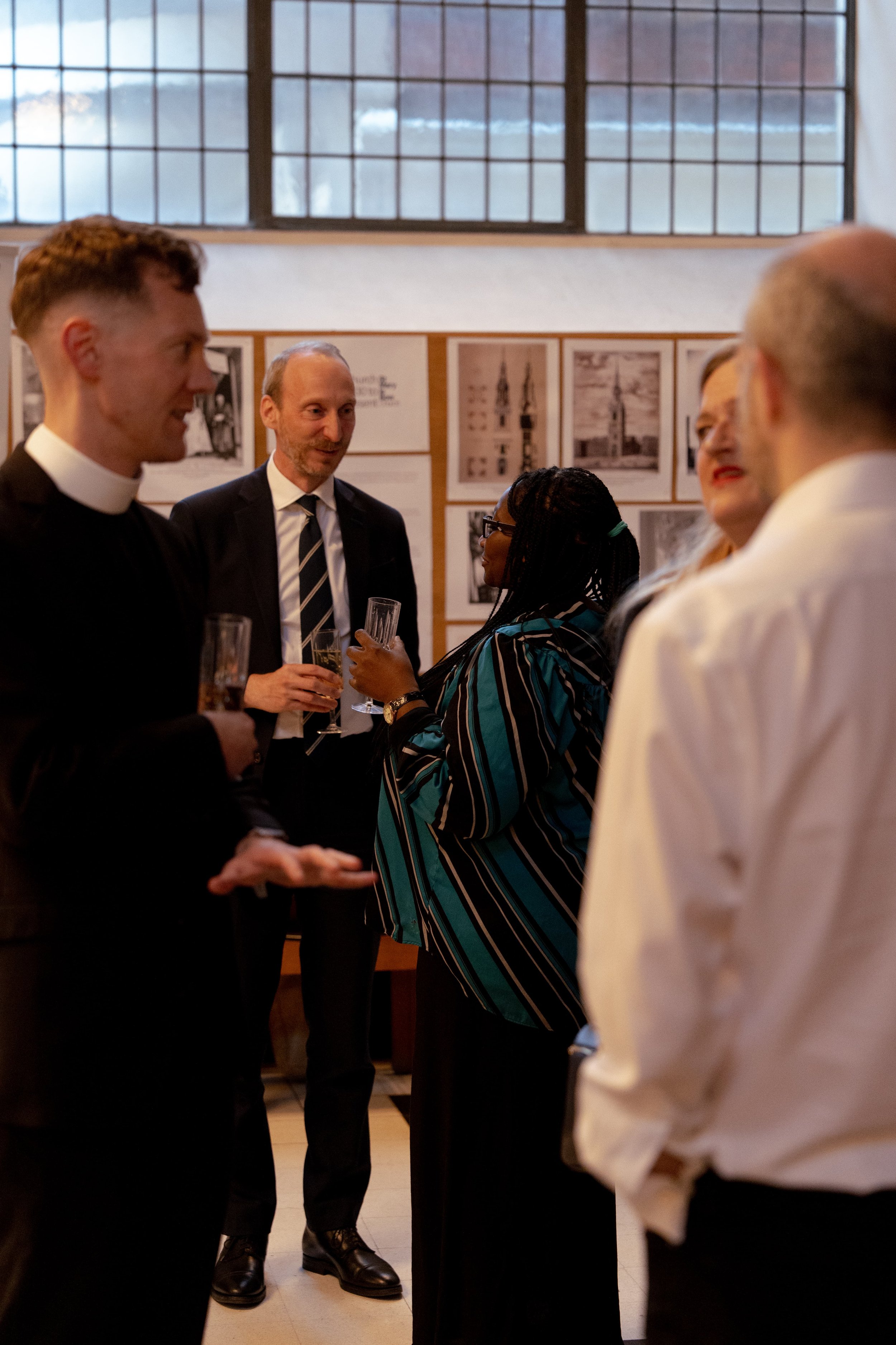 Group of people at a social gathering, dressed in formal and semi-formal attire, inside a room with large windows and framed pictures on the wall.