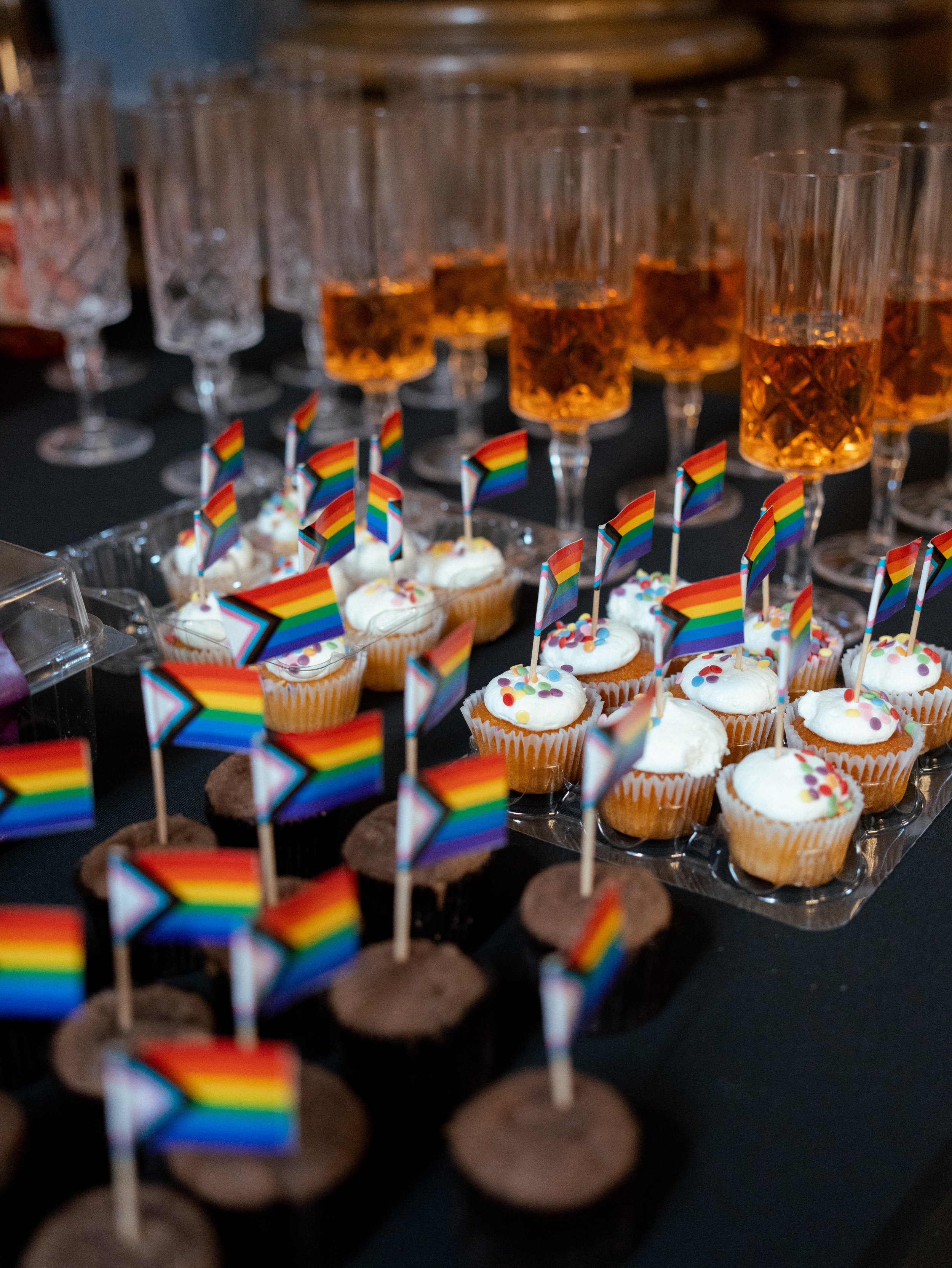 Assorted cupcakes with rainbow pride flags and colorful sprinkles, with glasses of amber-colored drinks in the background.