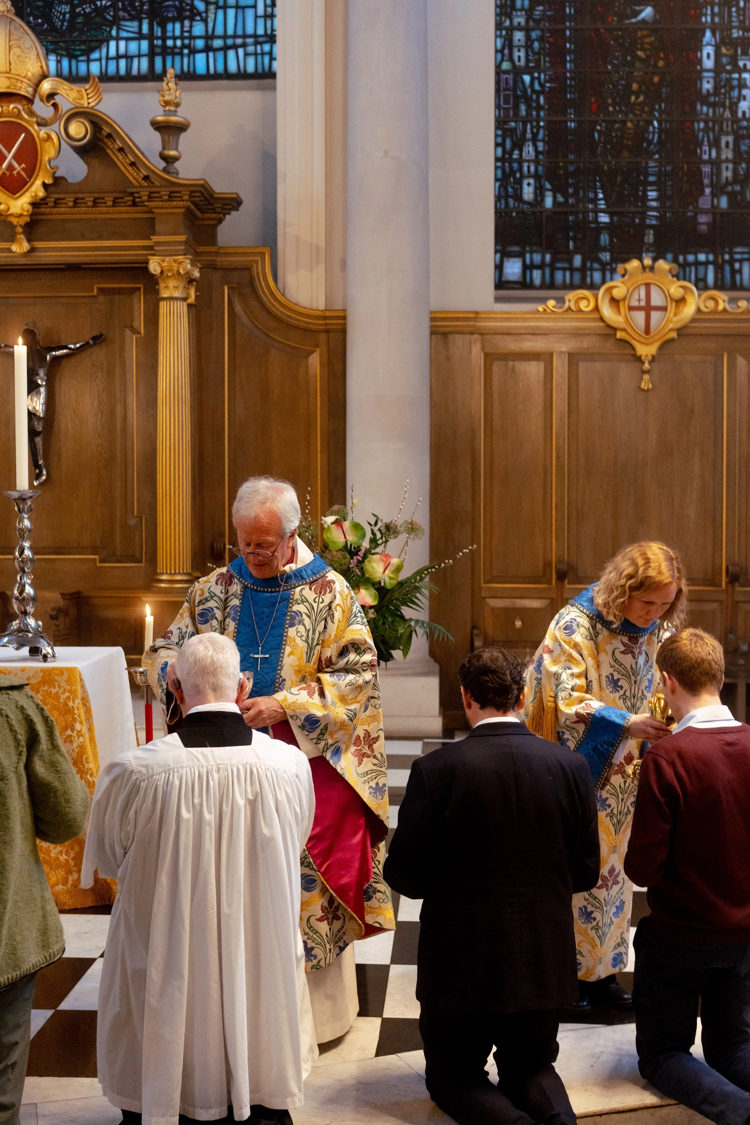 A religious ceremony in a church with clergy wearing ornate robes, individuals kneeling, and religious symbols such as a crucifix and candles.