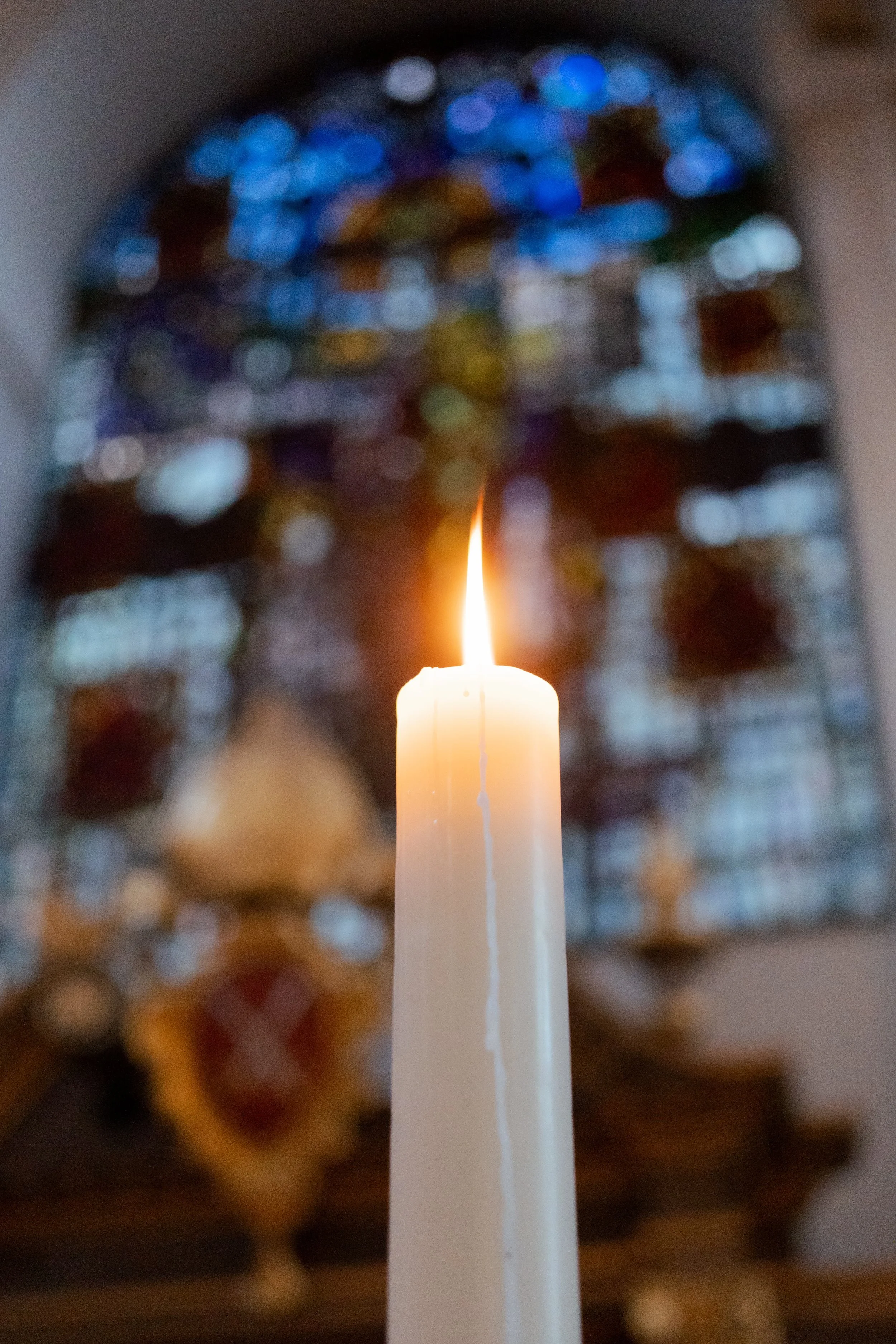 A lit white candle in front of blurred colorful Christmas ornaments or decorations.