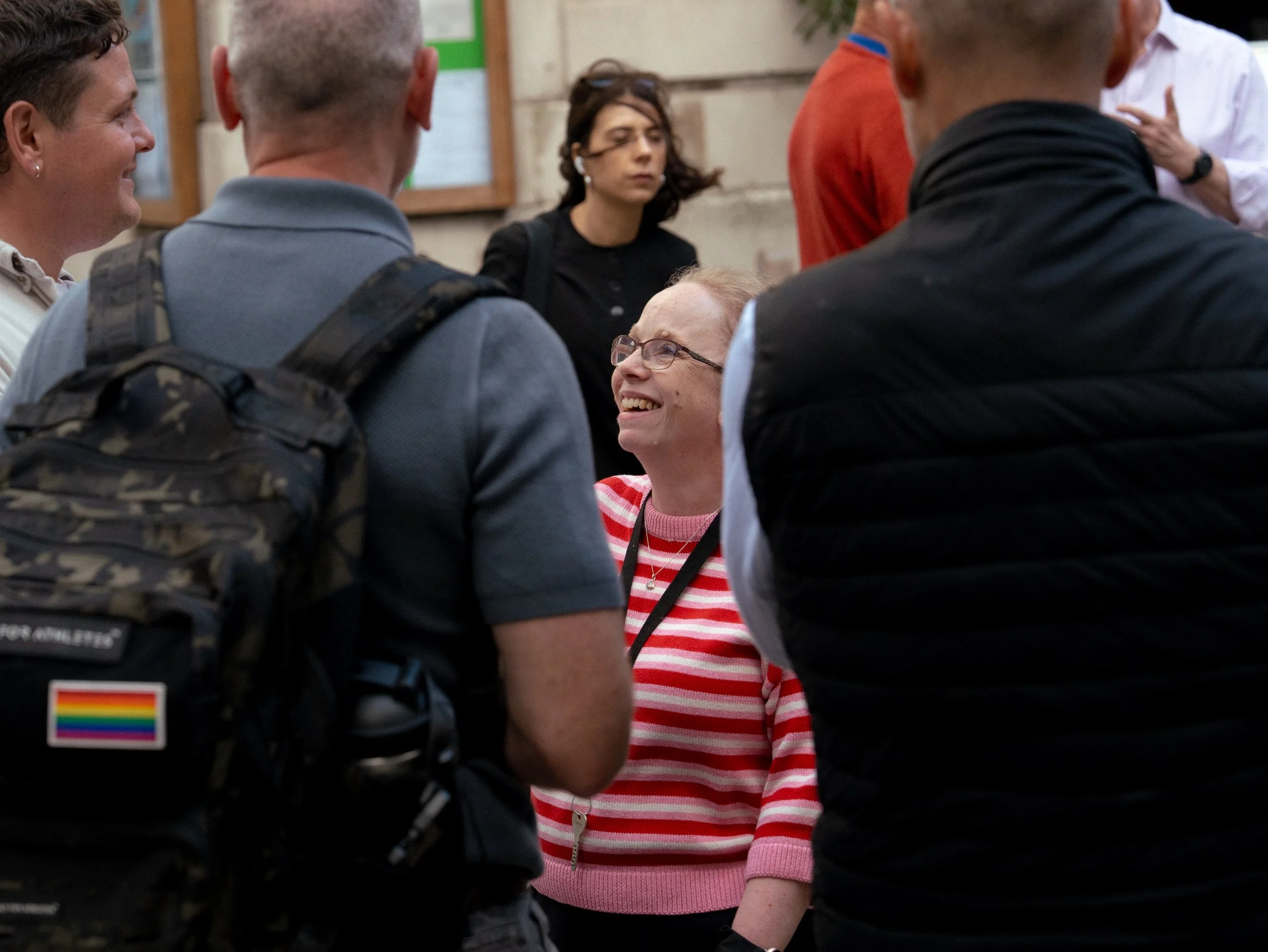 A group of people gathered around a smiling woman with glasses and a striped pink and white shirt, in an outdoor setting with a wall and bulletin board in the background. Others are standing and listening attentively.
