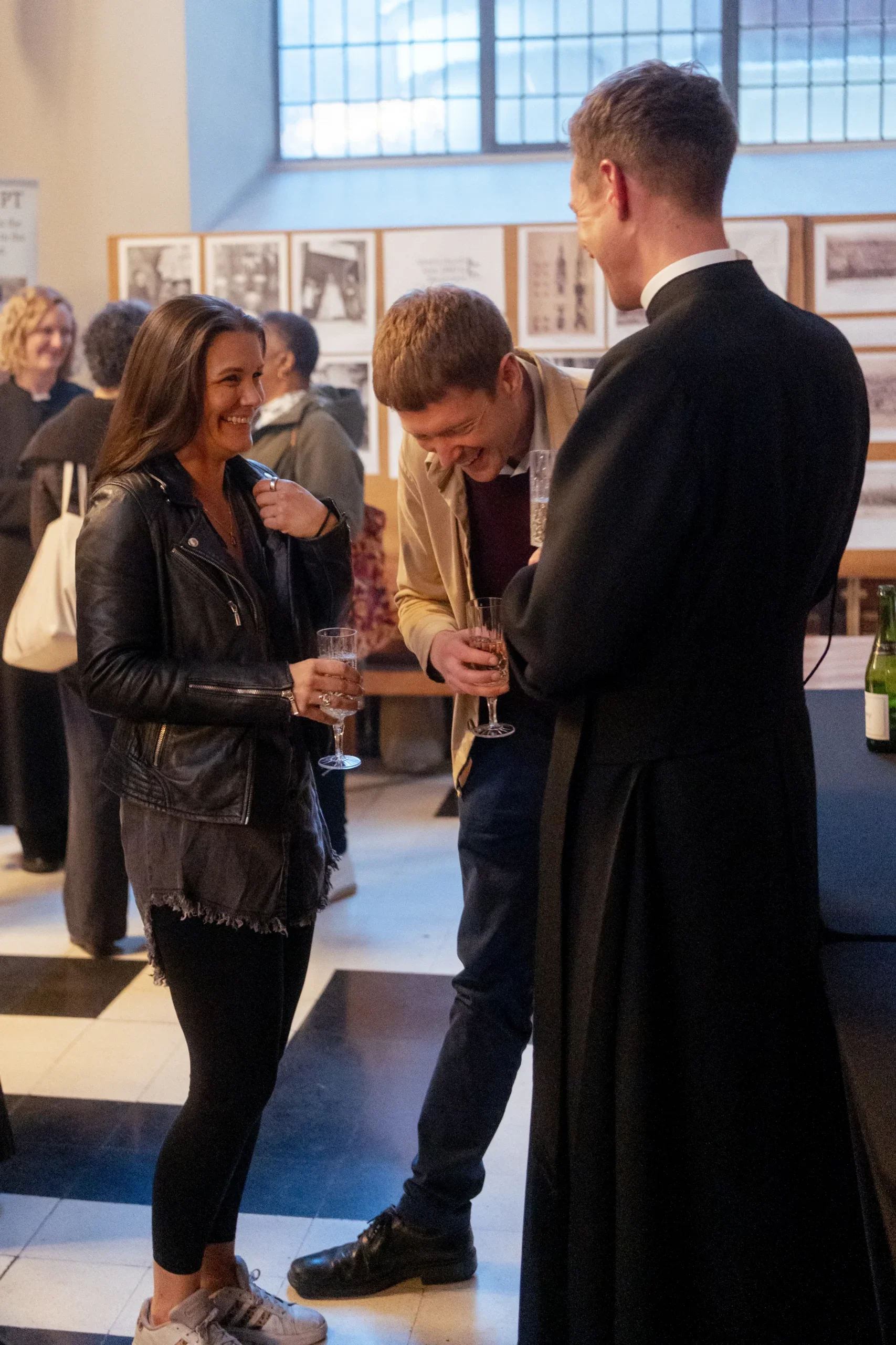 Four people are at a social event, with three of them engaged in a lively conversation. They are holding drinks and appear to be enjoying themselves, sharing a laugh. The setting looks like an indoor gallery or event space, with display boards and large windows in the background.