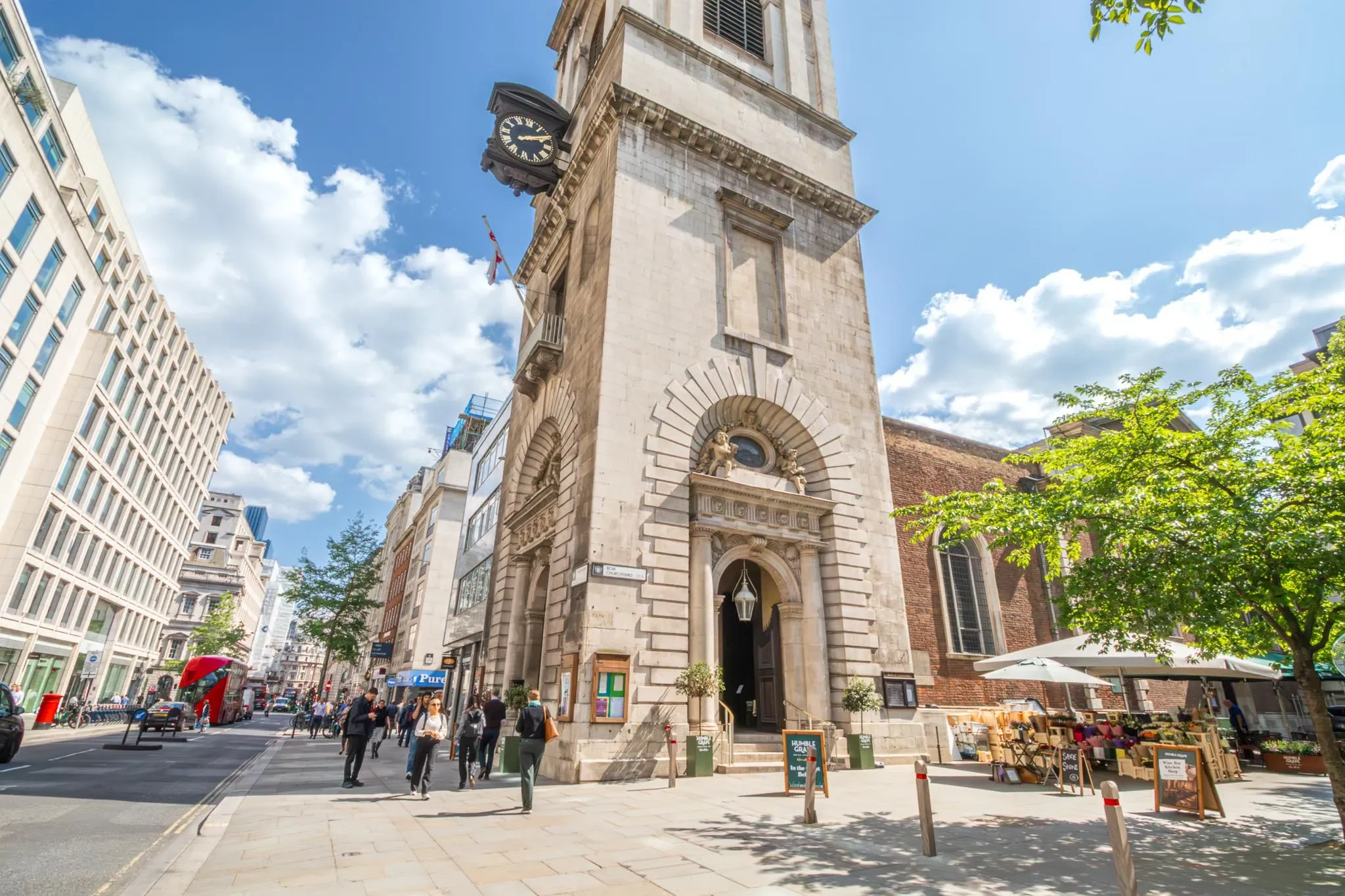 A busy city street in daytime with a historic clock tower at the intersection, surrounded by modern buildings and trees, with pedestrians, outdoor market stalls, and a double-decker bus.