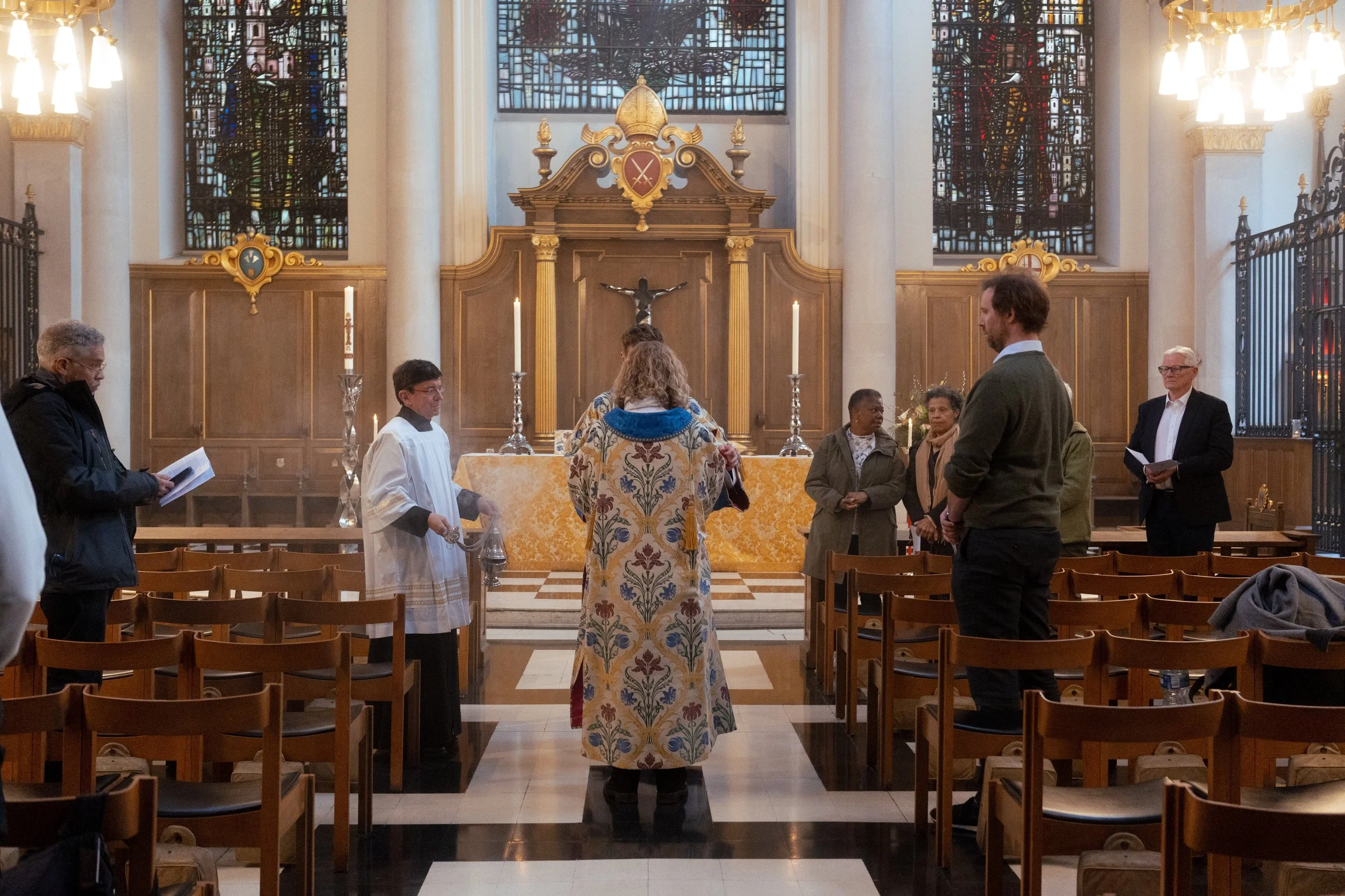 People gathered in a church during a religious service, with an altar in the background, stained glass windows, and lit candles.