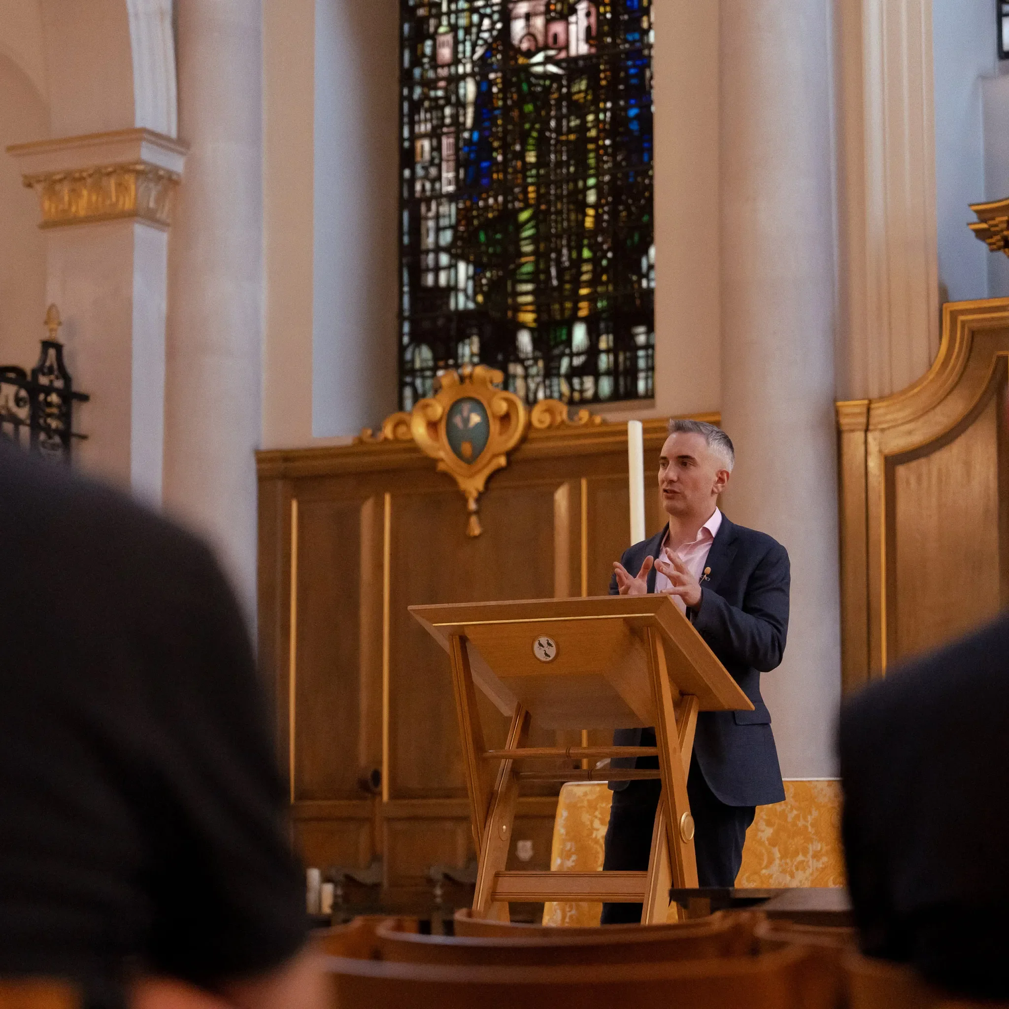 A man with gray hair wearing a dark suit and pink shirt, standing at a wooden podium in a church, speaking or giving a speech.