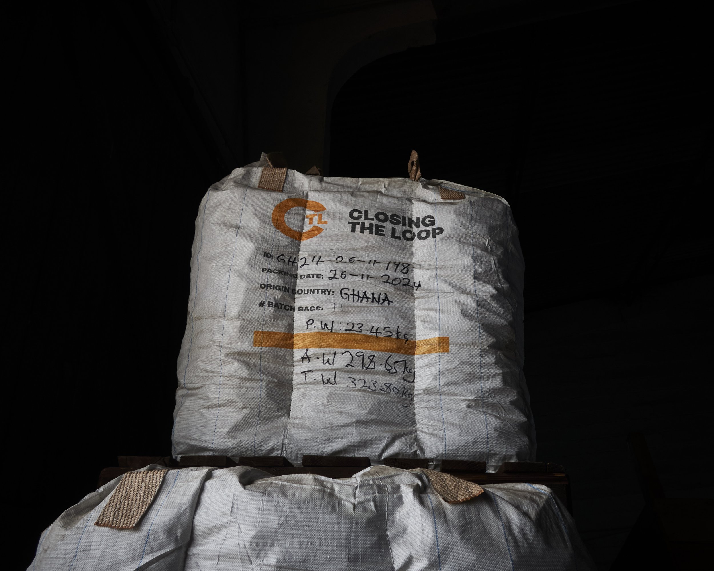A large white woven bag with orange and black text, contains handwritten information including origin from Ghana, packing date, and batch number, placed on a dark surface.