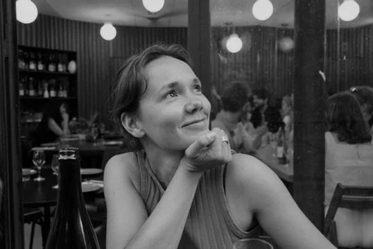 A woman sitting at a table in a restaurant, smiling and resting her chin on her hand, with other diners and hanging lights in the background.