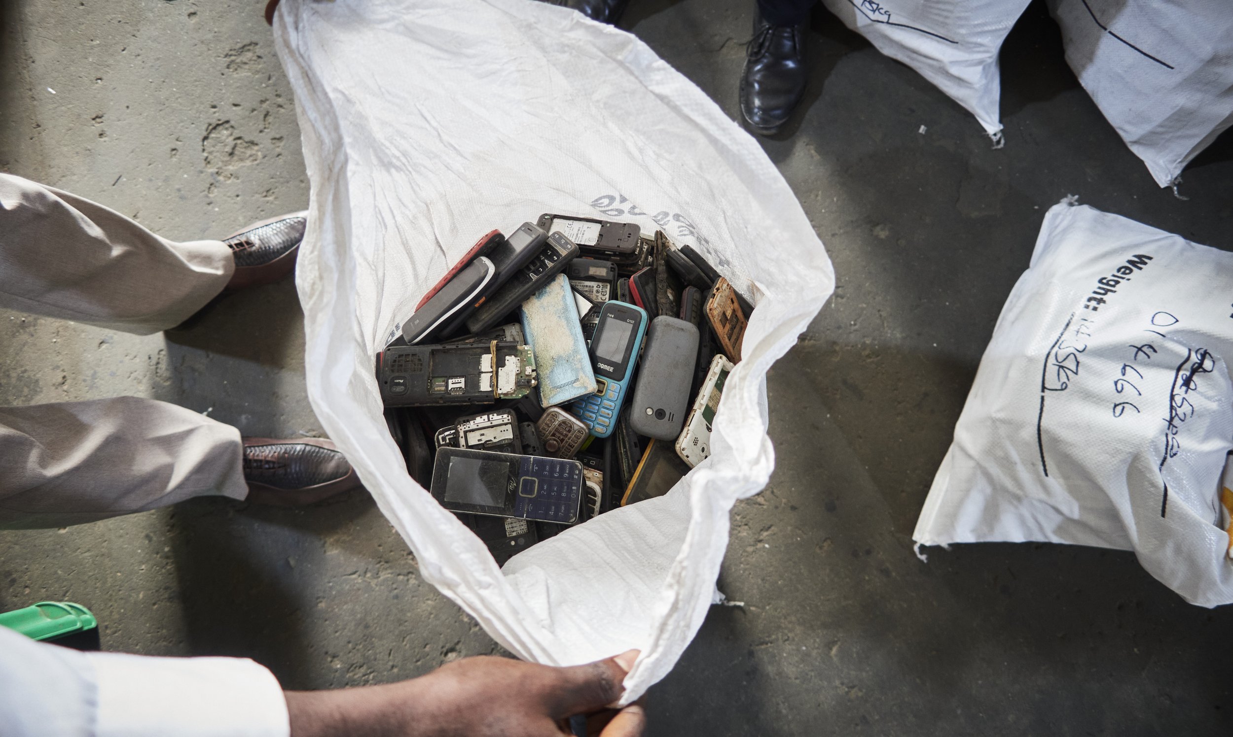 A large white plastic bag filled with old, disassembled cell phones and electronic devices on a concrete floor. Two people's legs and shoes are visible near the bag.