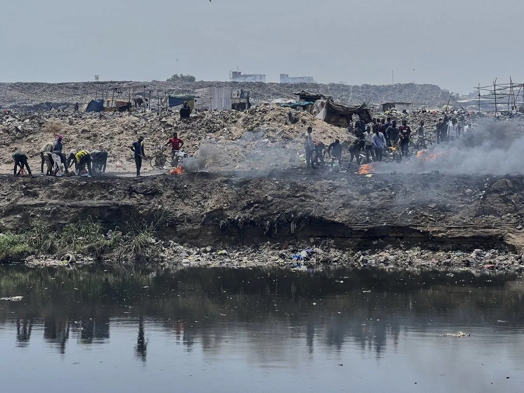 People gather near a landfill with smoke and small fires, with a body of water in the foreground and makeshift shelters in the background.