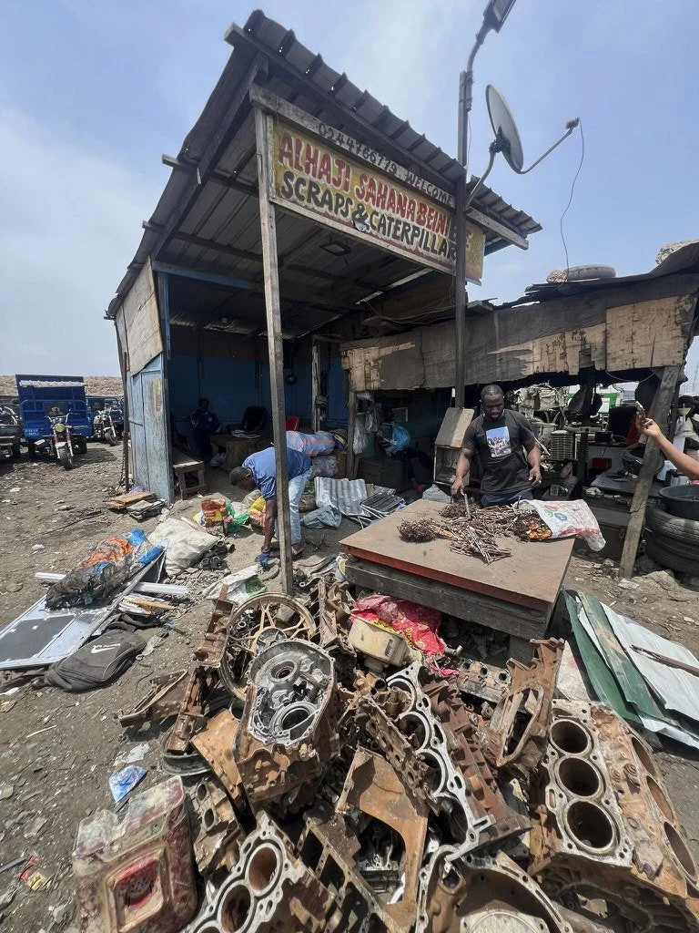 A small scrap and catering shop with a sign reading 'Alhaji Sahaba Ben Scrap & Caterpillar'. In front of the shop, there are rusty engine parts and debris. Several people are busy working nearby, with one person grilling on a makeshift grill. The are