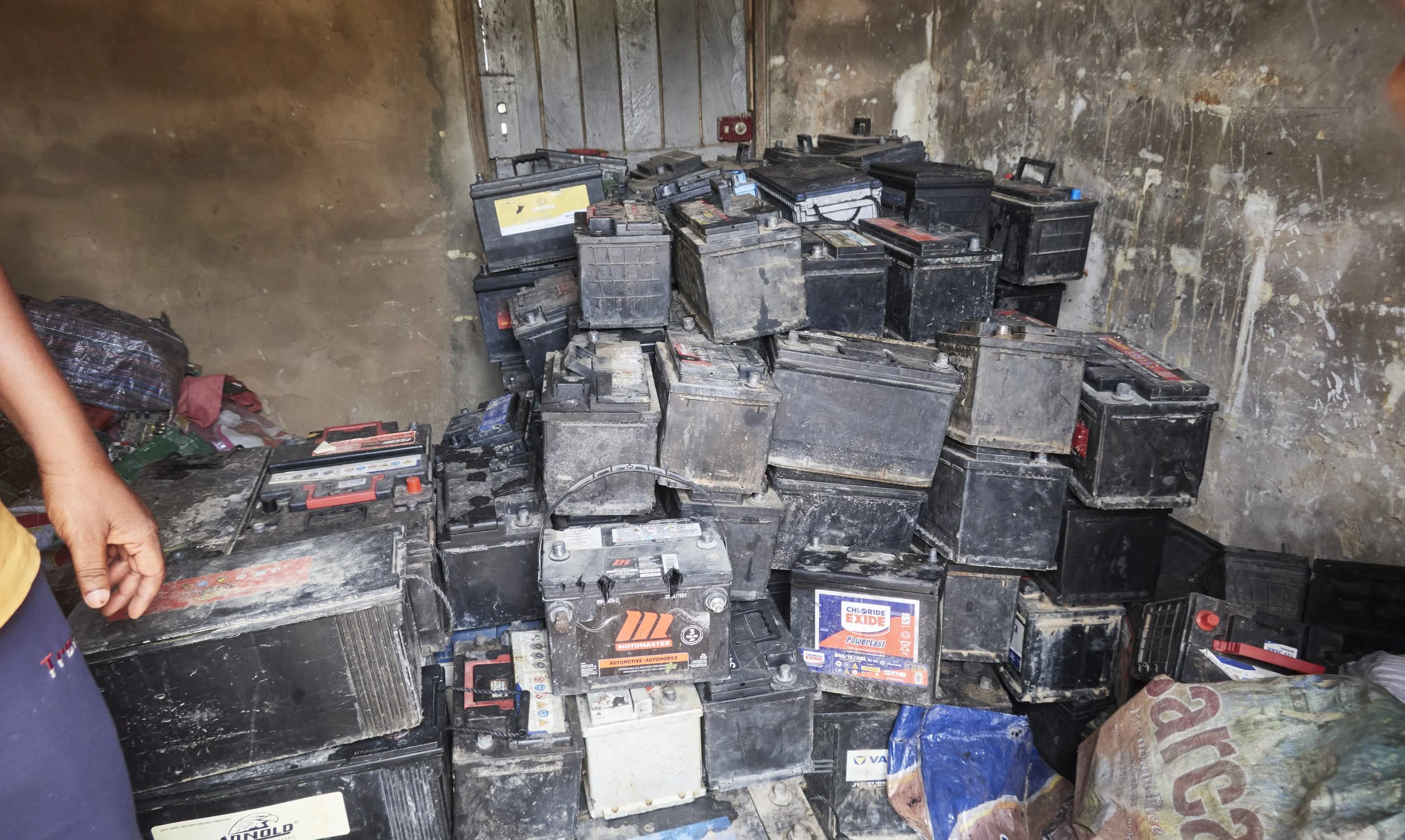 A large pile of old, dirty car batteries stacked on a rough concrete floor against a weathered wall.