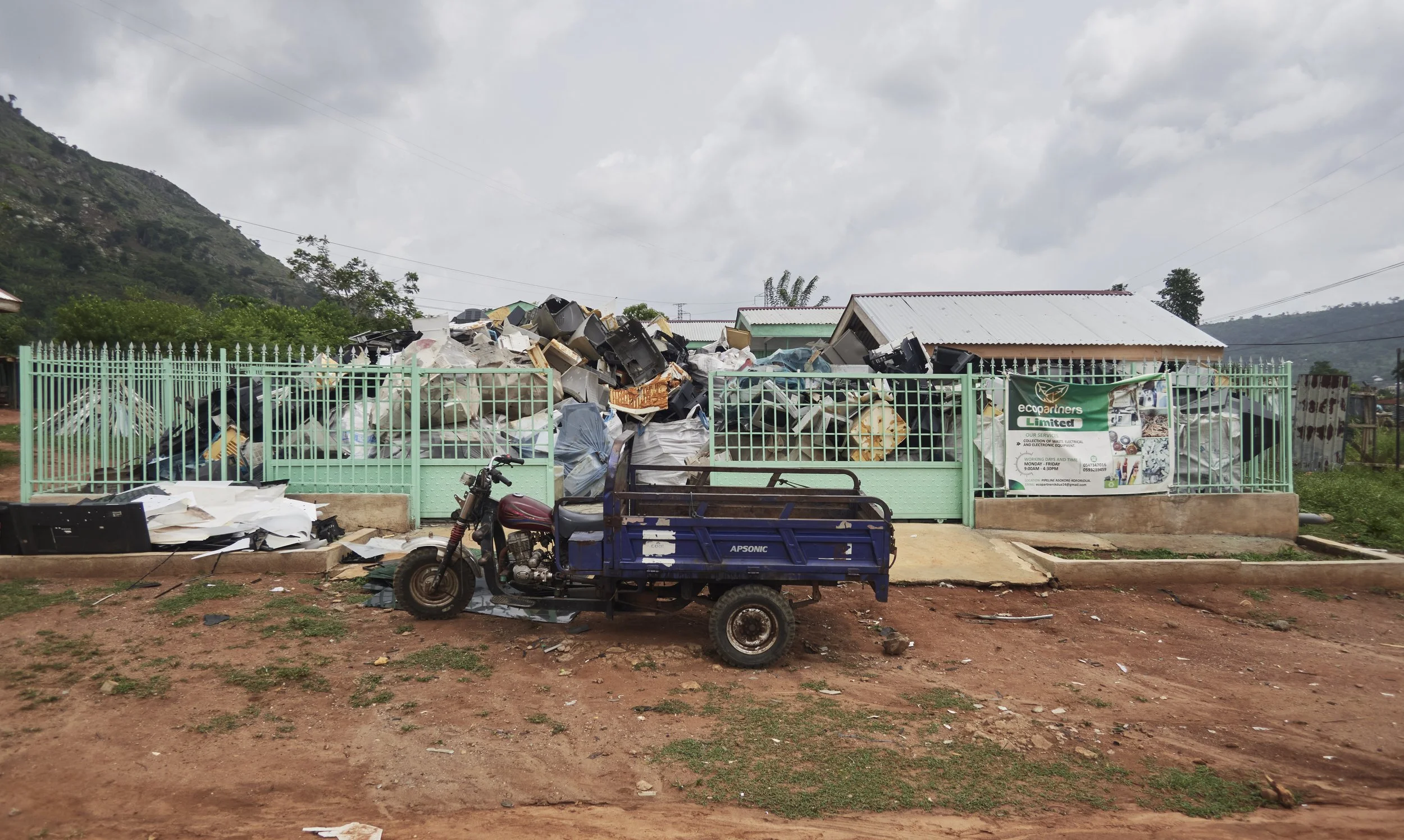 A green fence surrounds a large pile of discarded electronic waste, including old televisions, inside a facility with a small building, metal roof, and a motorcycle parked outside. Mountain hills can be seen in the background under a cloudy sky.