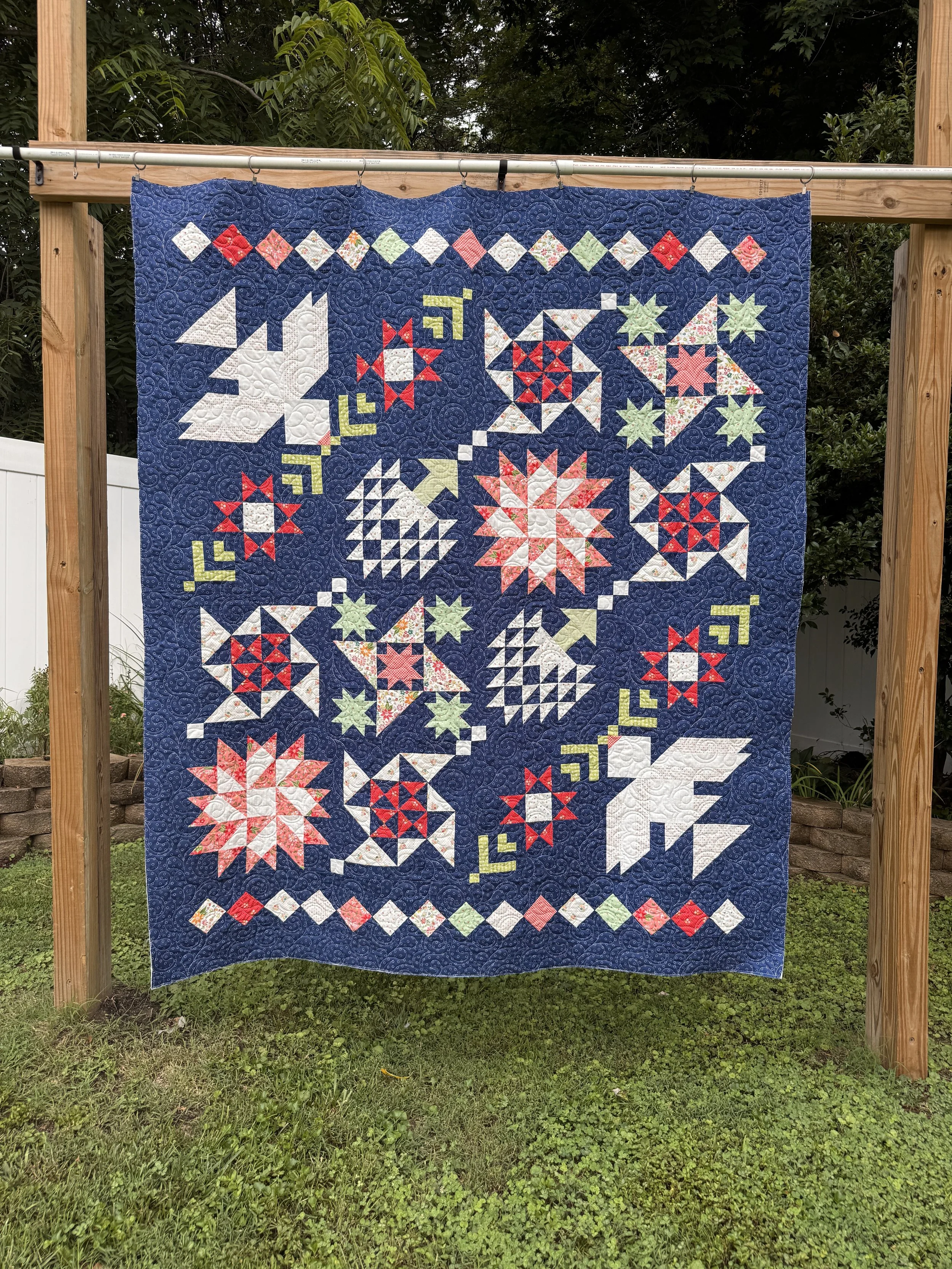 A completed quilt hanging on a clothesline outdoors, featuring intricate star and geometric patterns in red, white, green, and floral fabrics on a dark blue background.