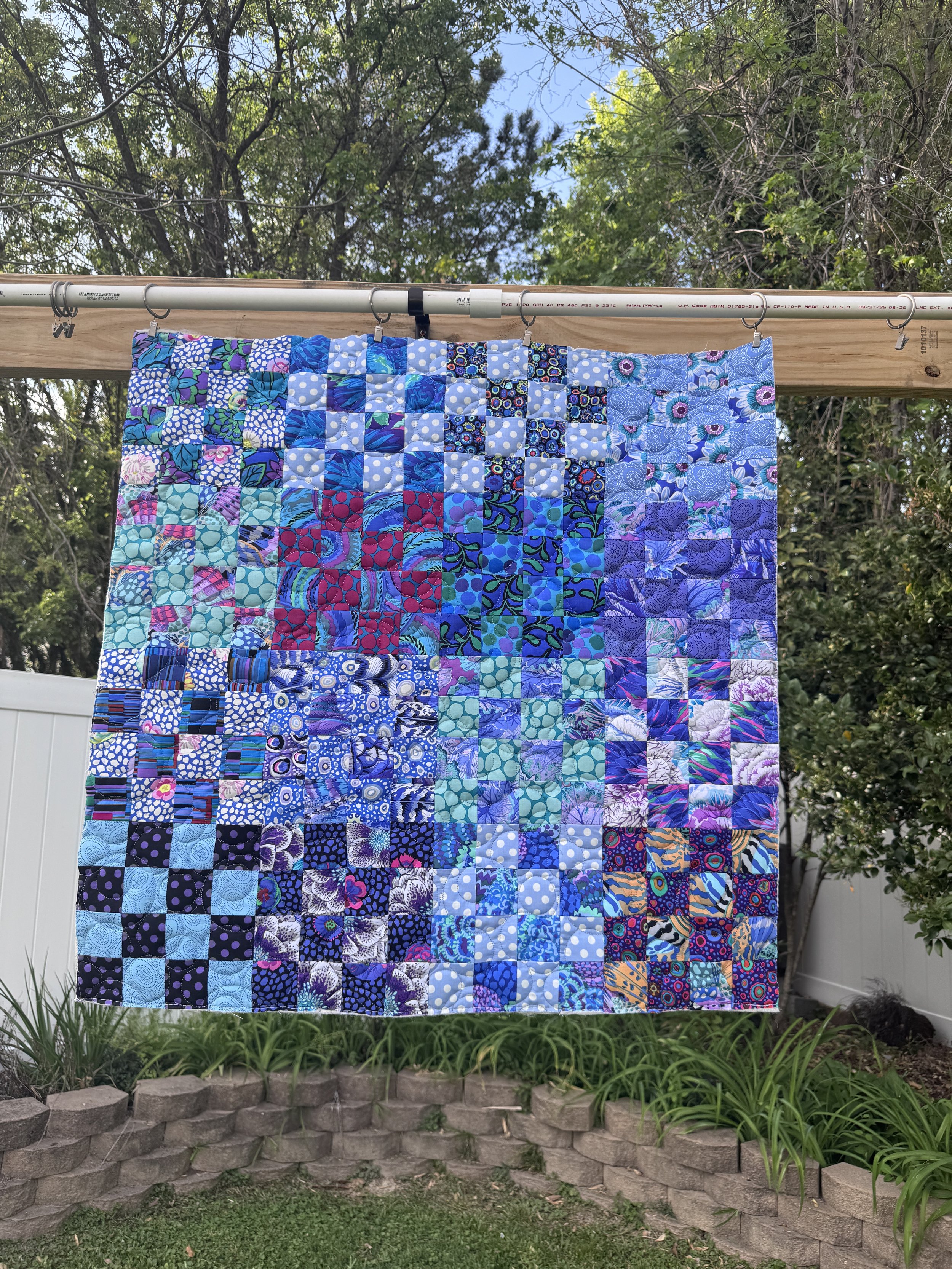 A quilt with a blue, purple, and pastel color pattern hangs on a clothesline in a backyard with trees, a white fence, a brick border, and green plants.