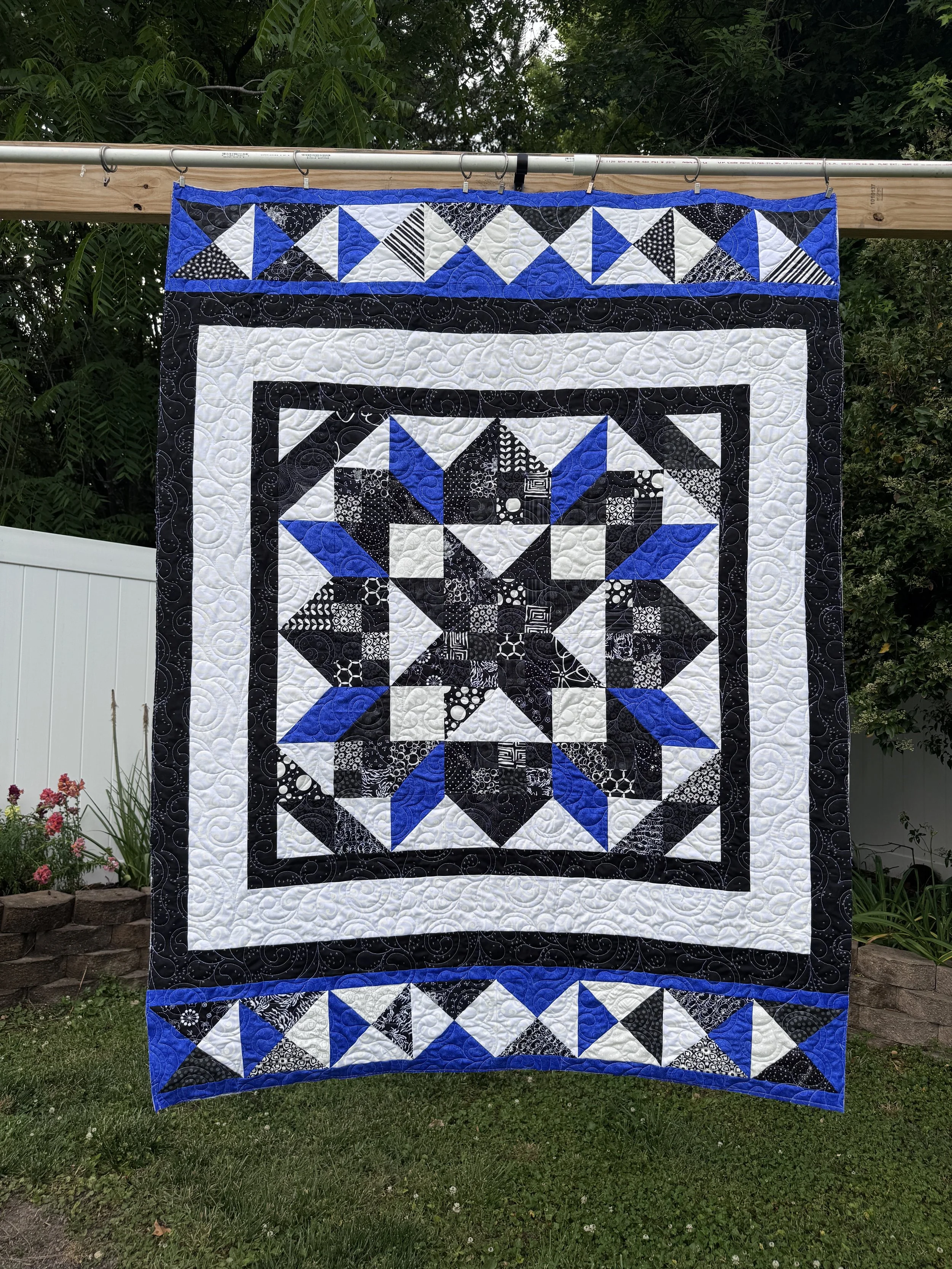 A quilt hanging on a clothesline outdoors, featuring a pattern of blue, black, and white geometric shapes, with intricate stitching, surrounded by greenery and flowers.