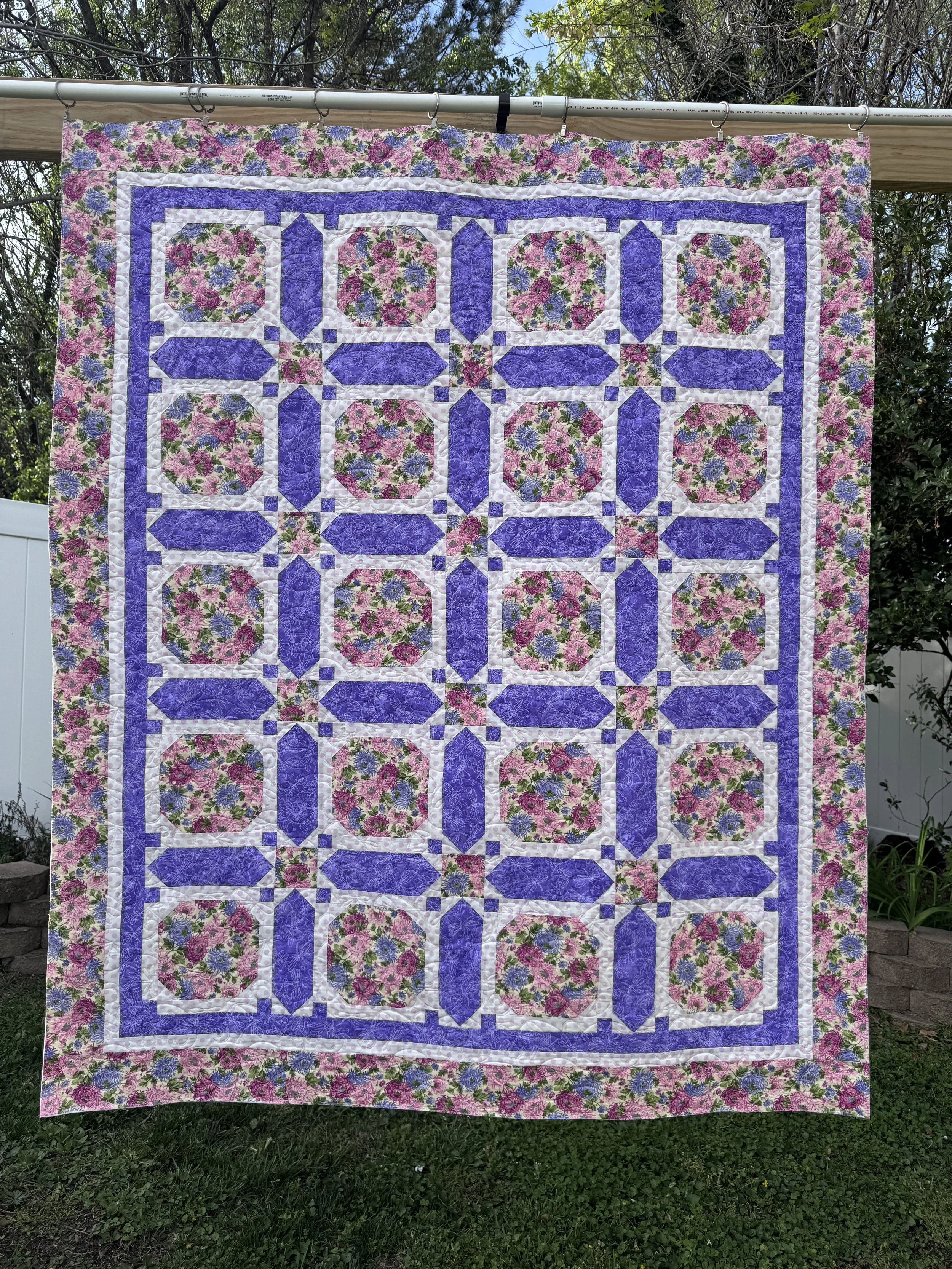 A colorful patchwork quilt hanging outdoors on a clothing line in a garden with trees and grass background.