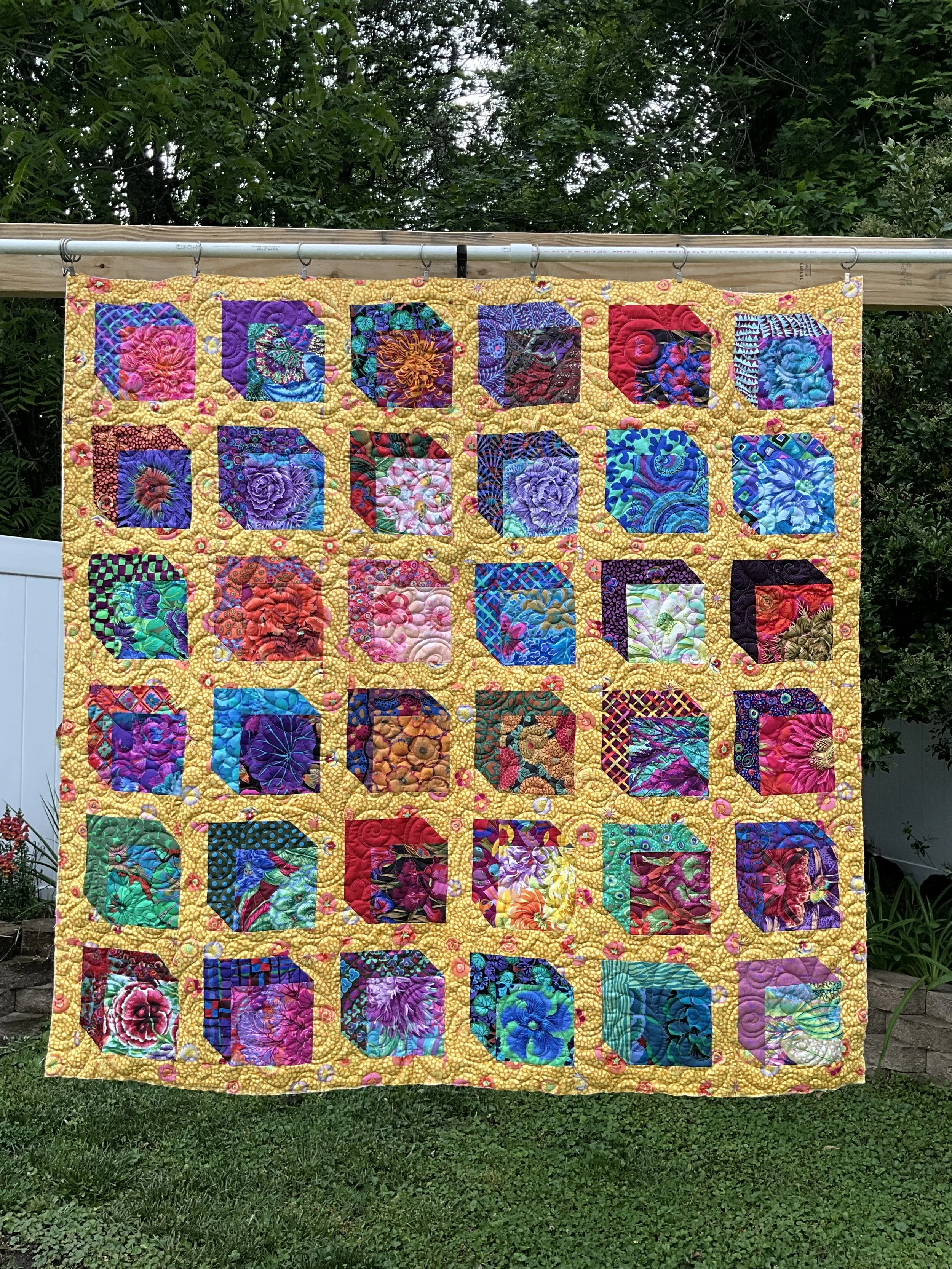 A colorful quilt hanging outdoors on a clothesline, with leafy green trees in the background and grass below.