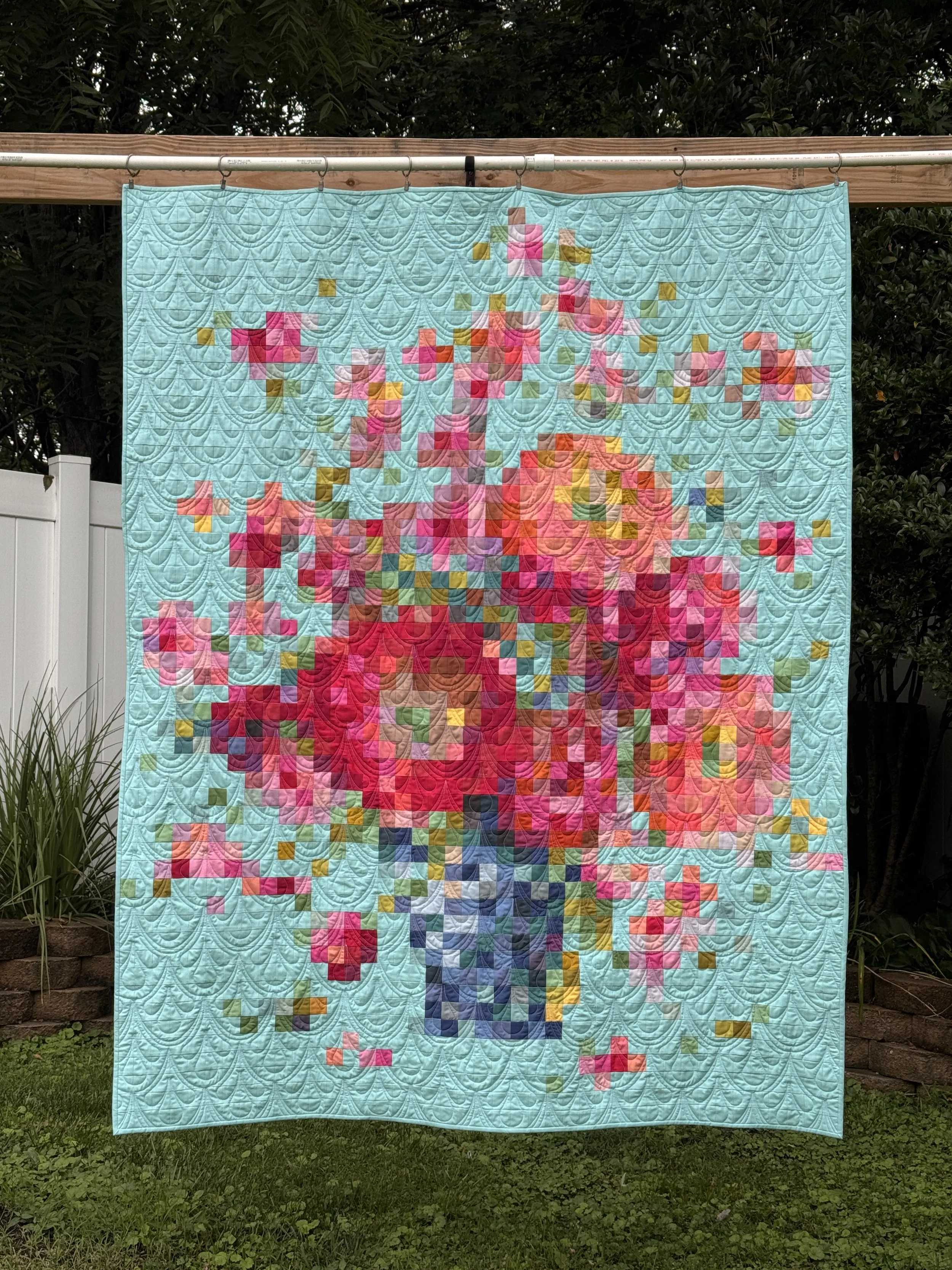 Colorful quilt with pixel art design of a pink and orange sunset over green hills, mounted on a clothesline outdoors.