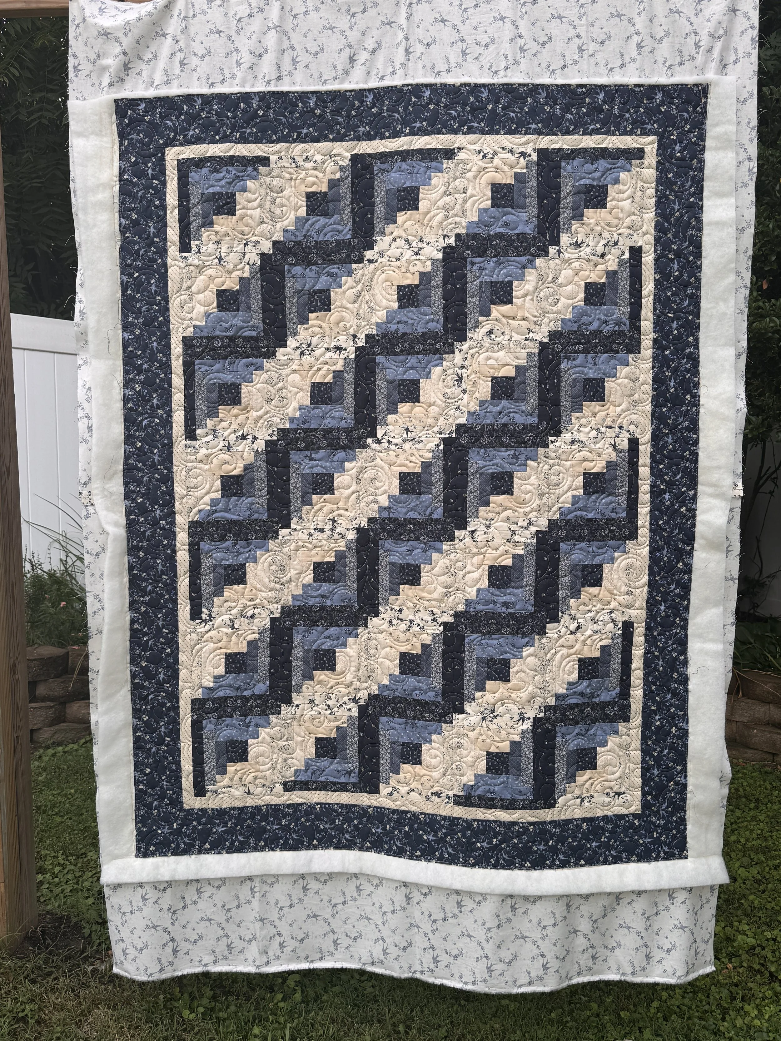 A quilt hanging outdoors on a clothesline with a white floral border and a dark blue and beige pattern in the center.