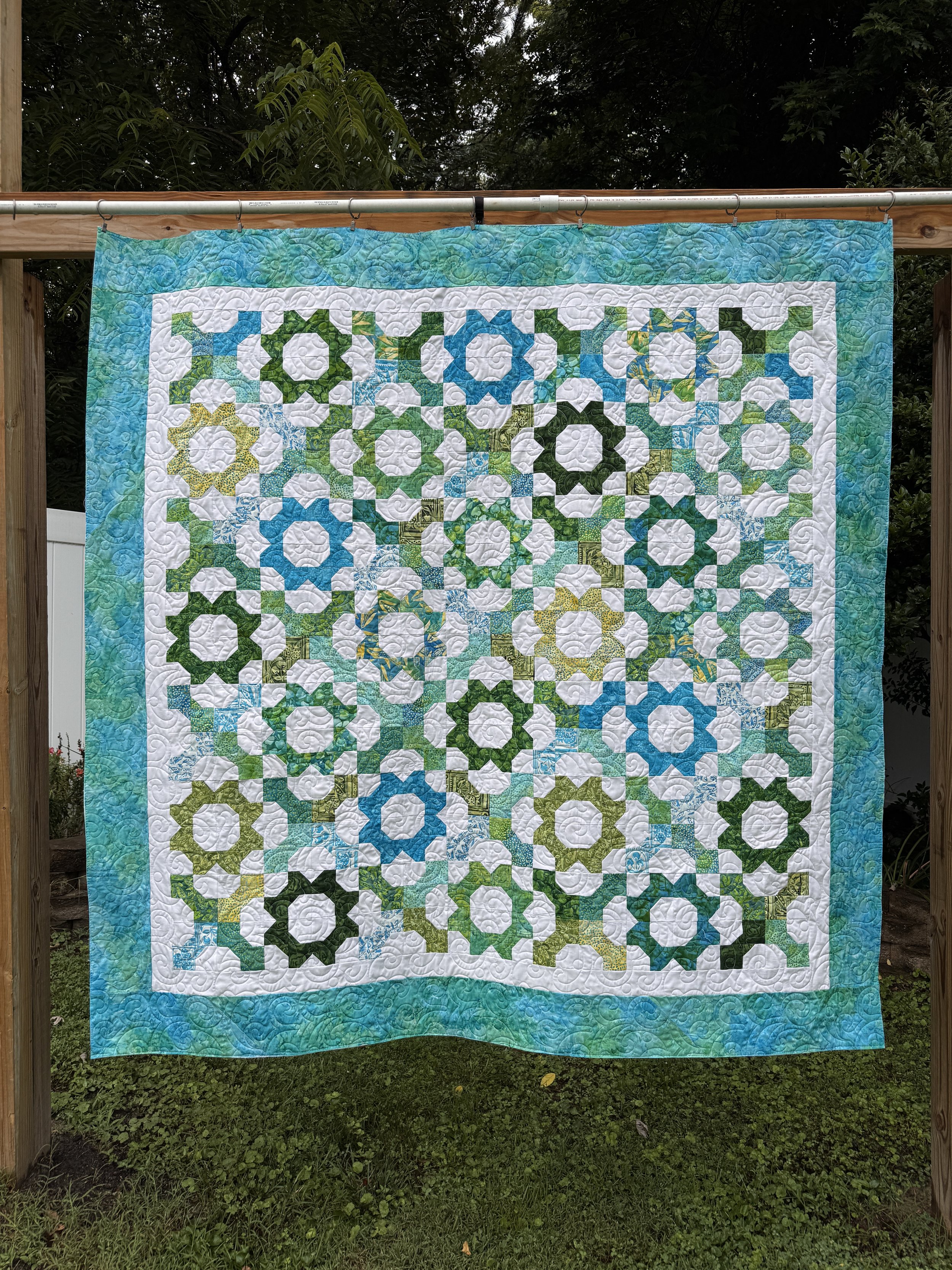 Colorful quilt with intricate patterns hanging outdoors against a backdrop of trees and greenery.