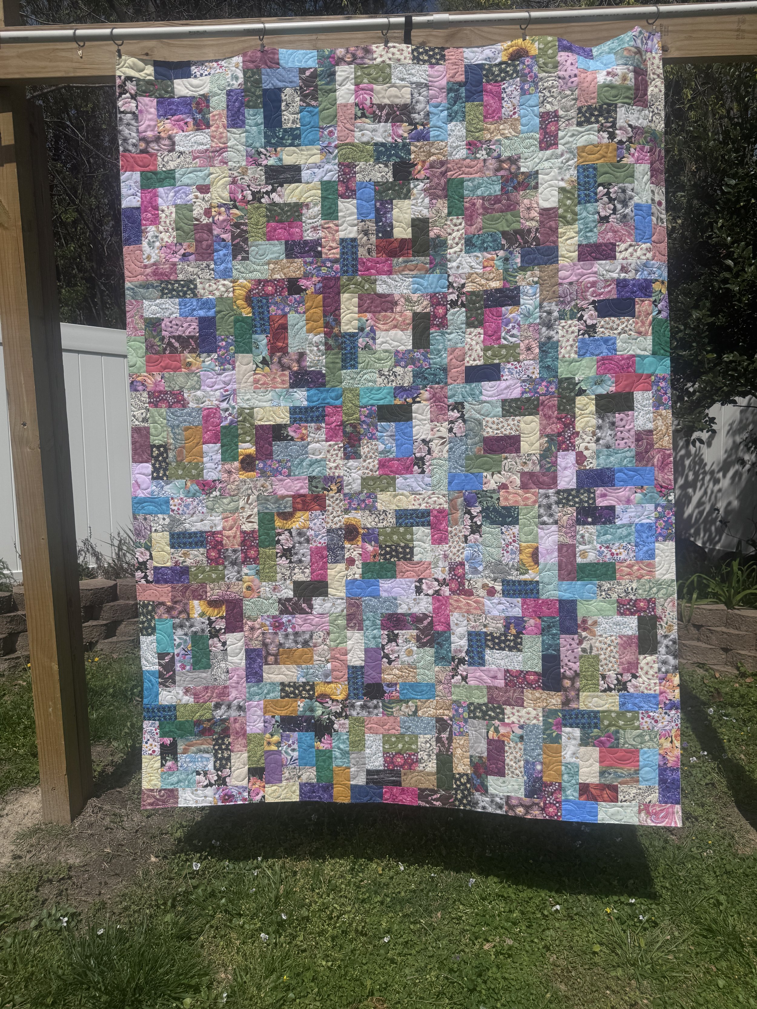 A colorful quilt with various floral and patterned fabrics hanging outdoors on a wooden frame.