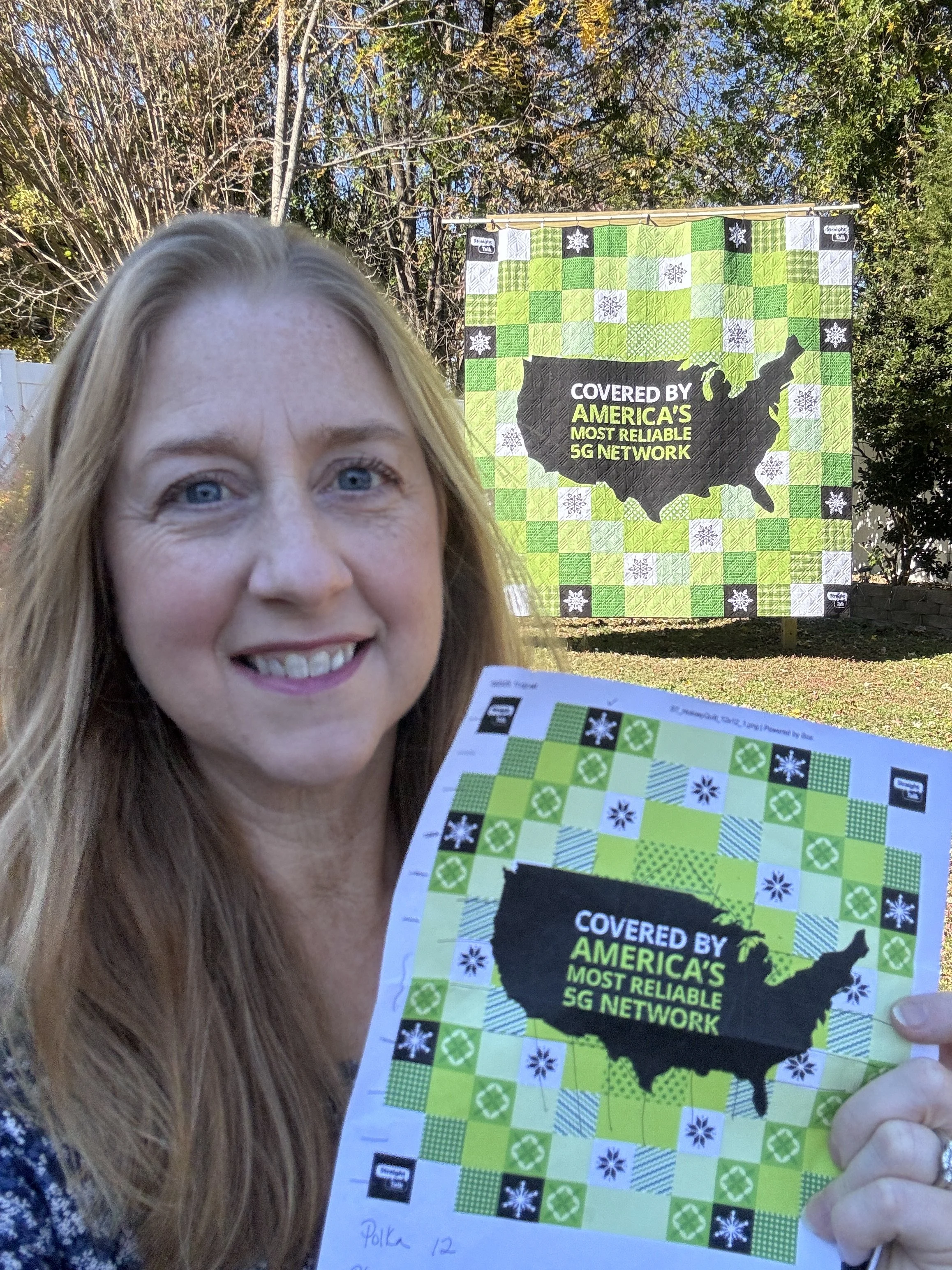 A woman with long light brown hair and blue eyes smiling outdoors, holding a colorful printed sheet. In the background, there is a large quilt or banner with a black map outline of the United States, surrounded by a patchwork of green, white, and black squares with snowflake patterns, and the text 'Covered by America's Most Reliable 5G Network'.
