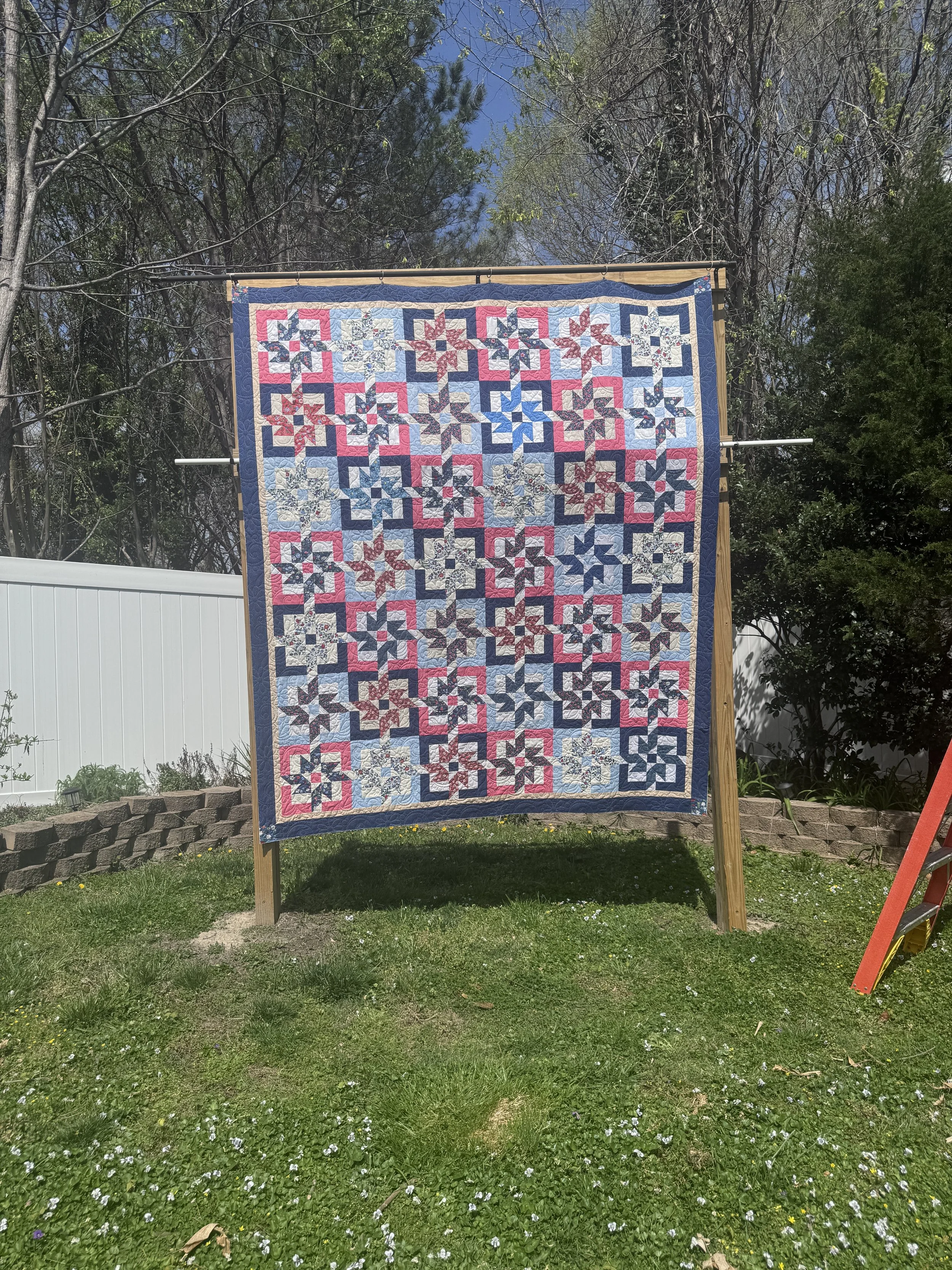 A colorful quilt hanging on a wooden frame in a backyard, with trees and a white fence in the background, and a green grassy area with small white flowers in the foreground.