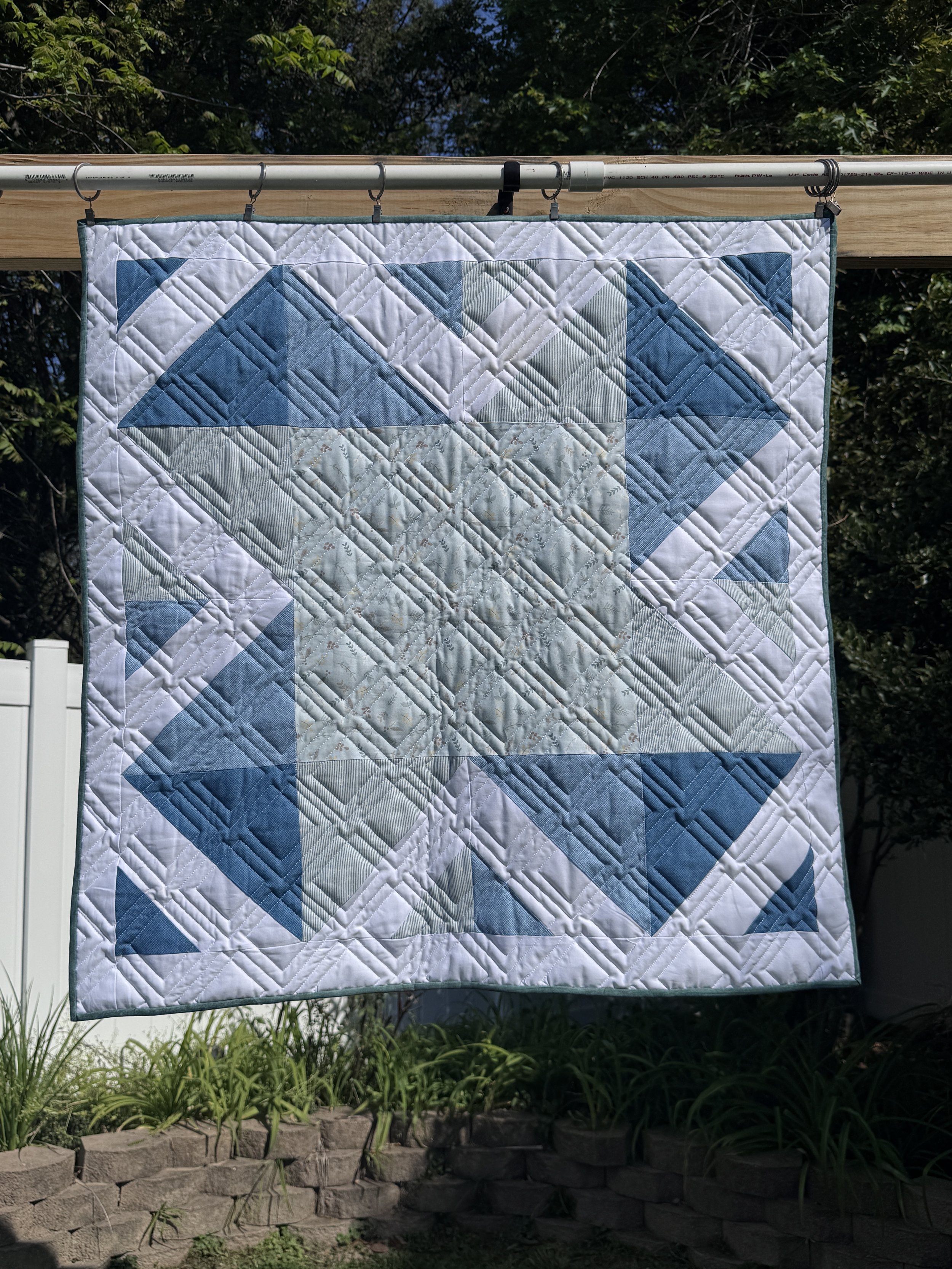 A quilt hanging outdoors on a clothesline, showing a geometric pattern in shades of blue, white, and beige, with a background of trees and a brick border.