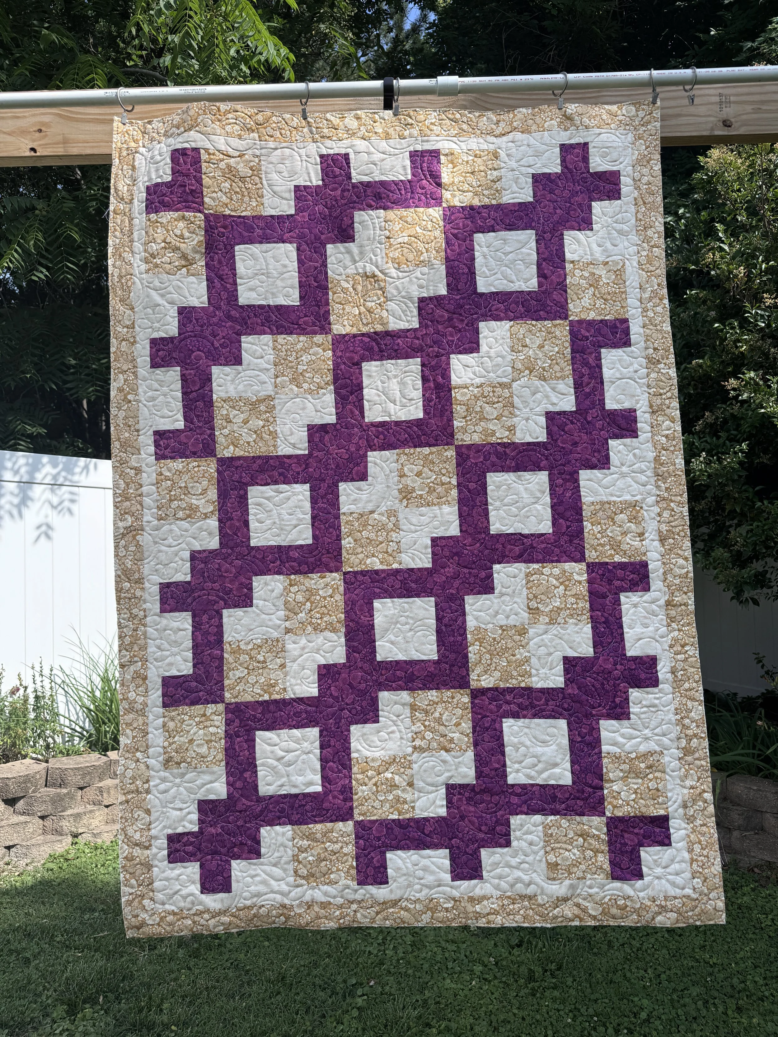 A colorful quilt hanging outdoors on a wooden pole, featuring a pattern of purple and tan blocks with a floral border, surrounded by greenery.