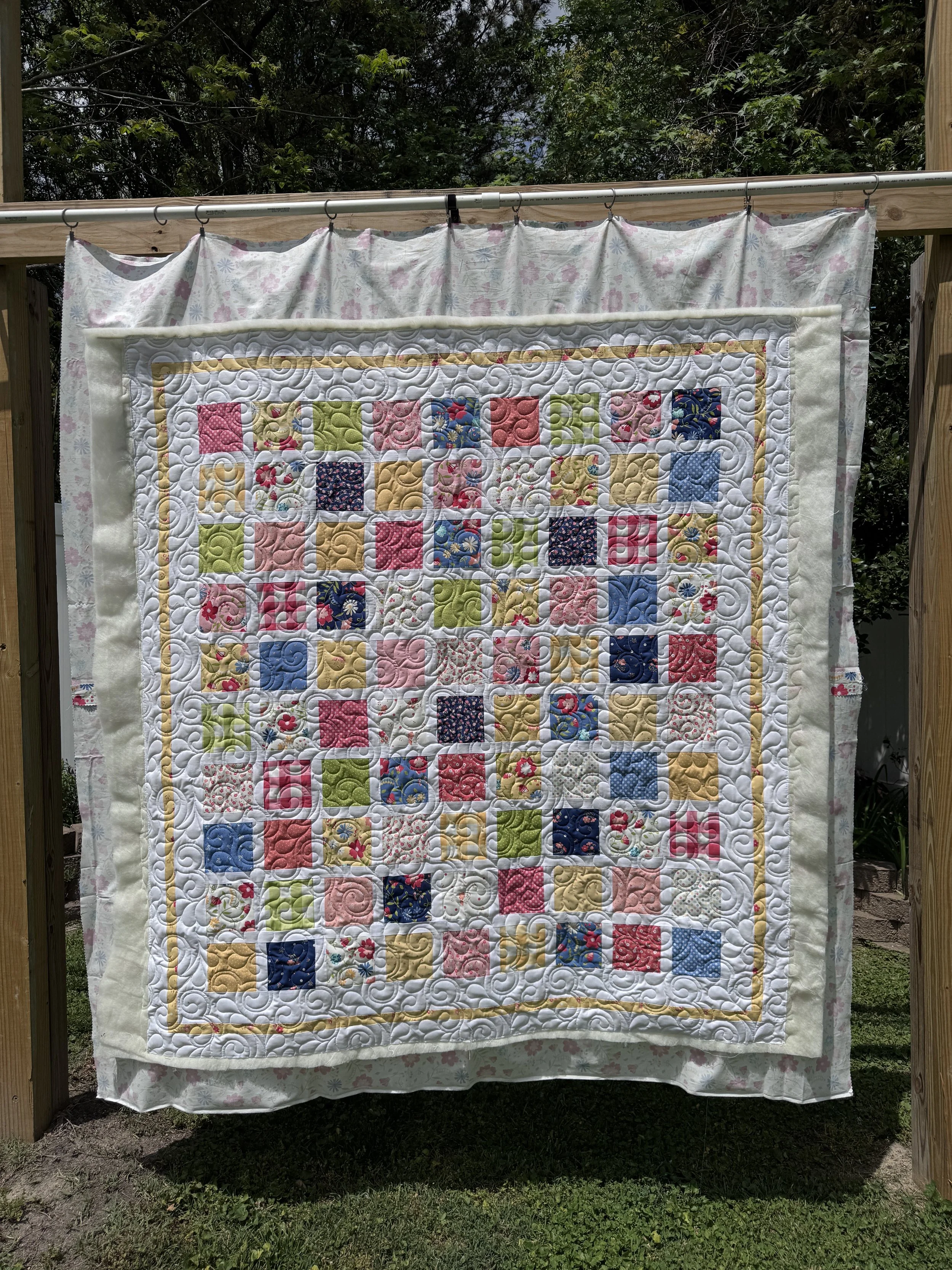 Colorful quilt with various patchwork squares hanging on a clothesline outdoors, with a floral fabric backdrop and wooden posts.